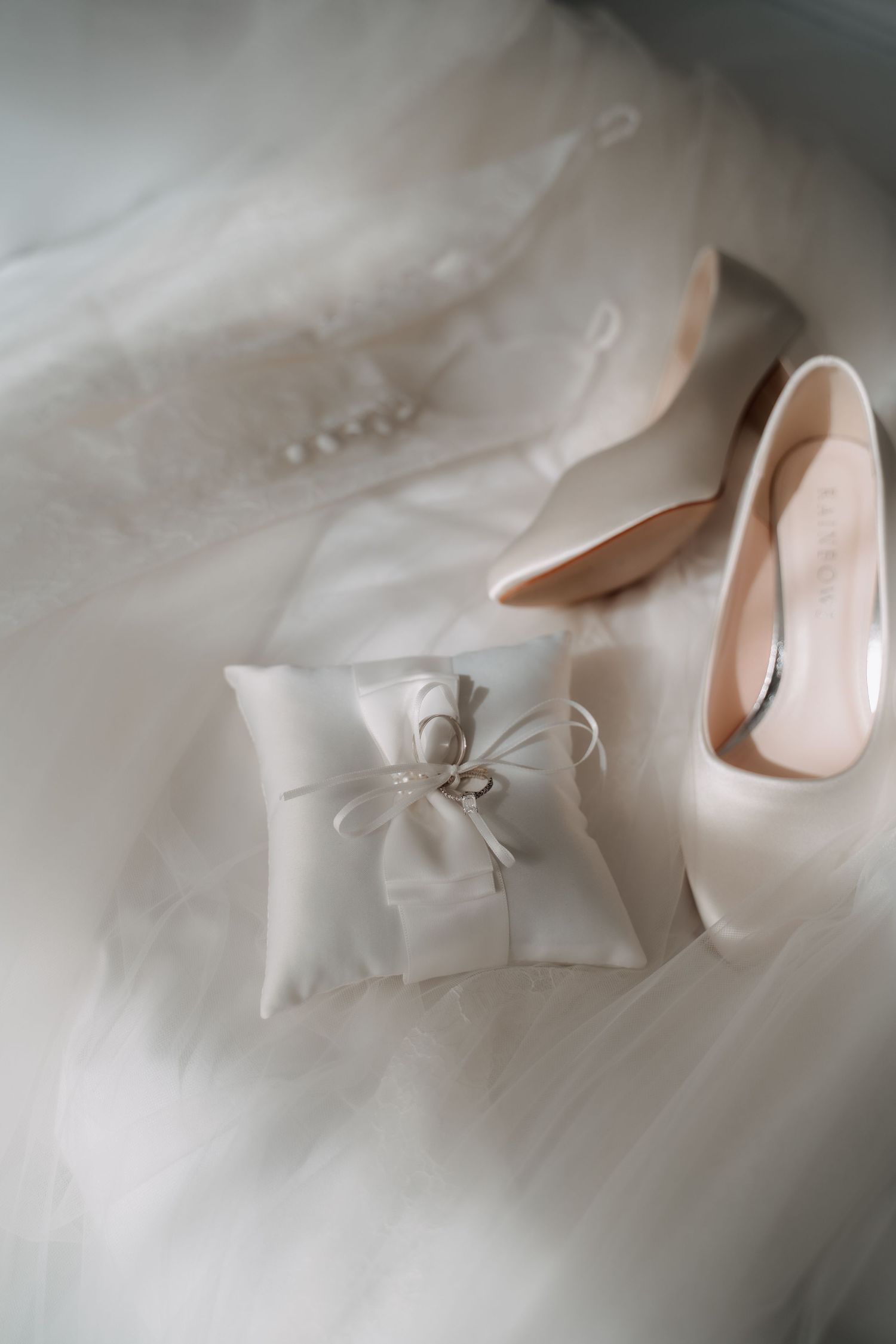 Wedding rings on white satin pillow beside nude heels and bridal gown, elegant bridal accessories flat lay.