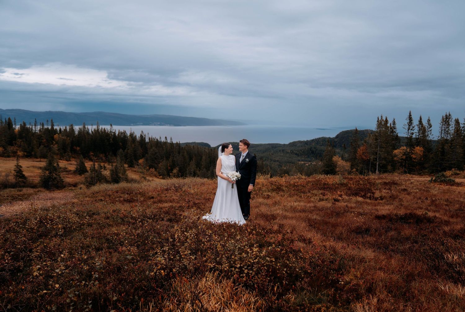Bride and groom embrace on a scenic hilltop with autumn foliage, evergreen trees, and a dramatic cloudy coastal backdrop.