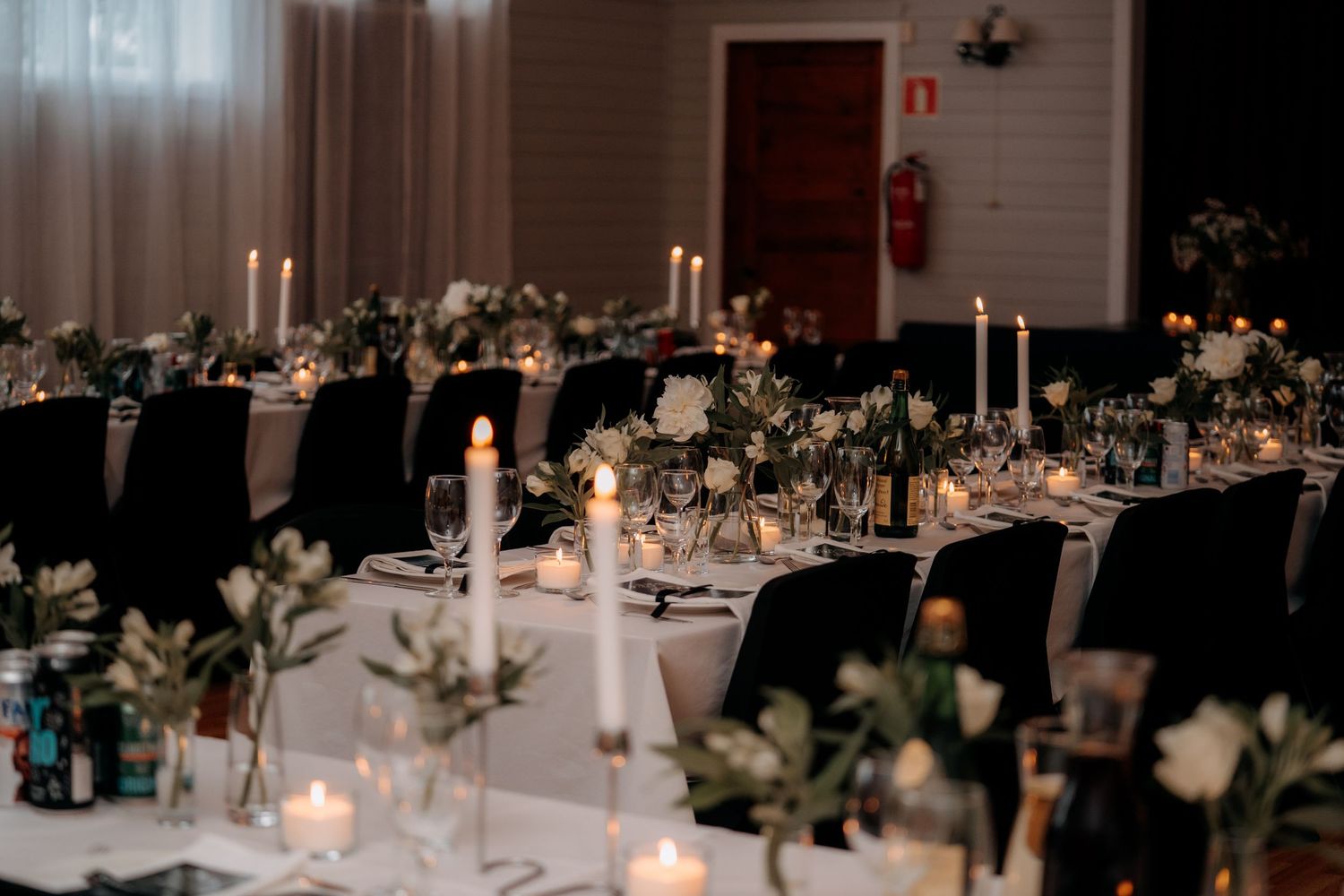 Elegant wedding reception hall with white tablecloths, black chair covers, tall white candles, and floral centerpieces.