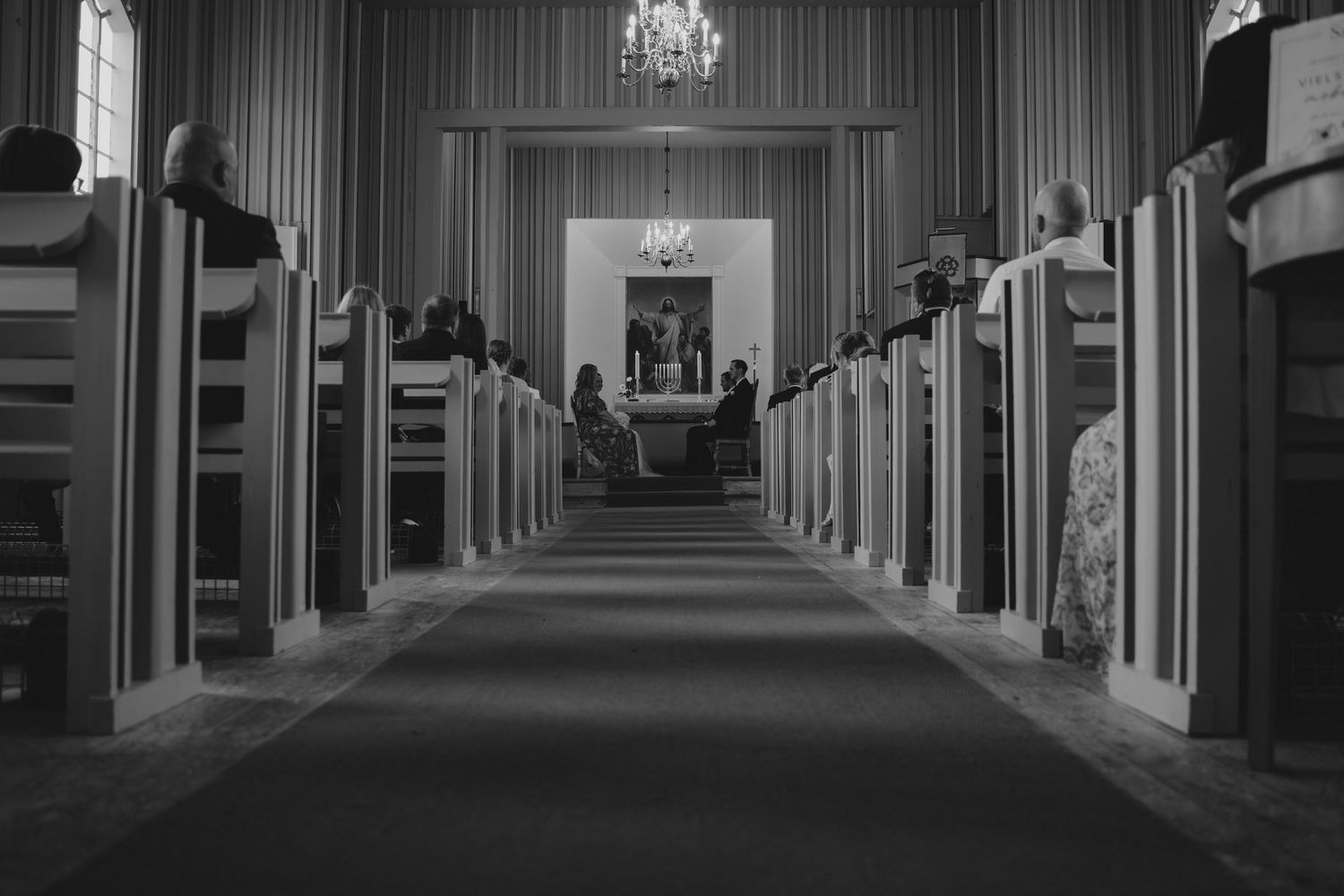 Black and white photo of a church aisle with wooden pews leading to an altar, chandelier overhead.