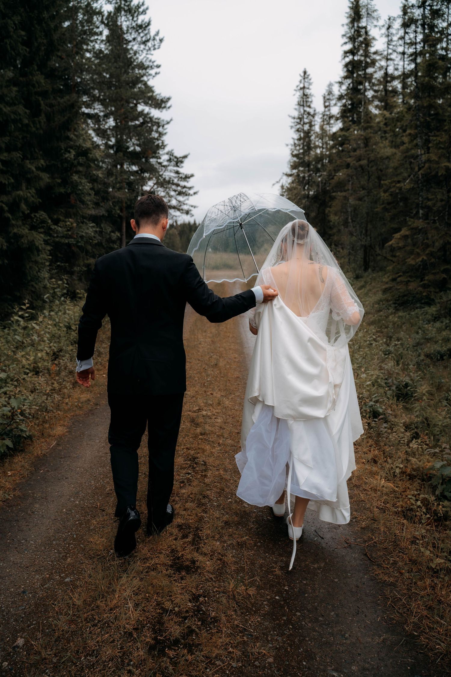 A bride and groom walk hand-in-hand down a forest path in the rain, sharing a clear umbrella on their wedding day.