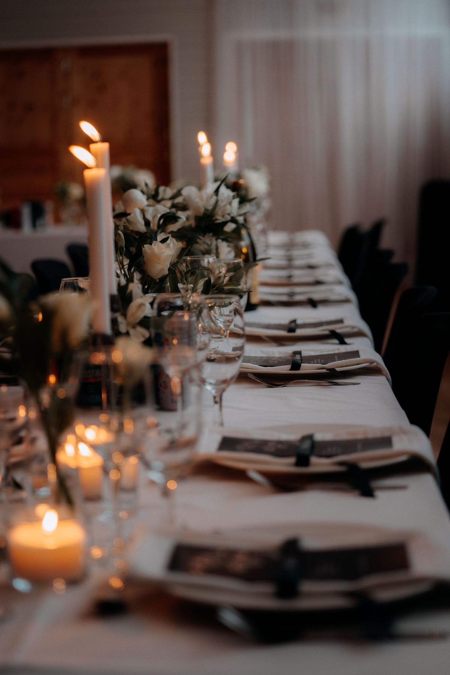 Elegant candlelit reception table with white floral centerpieces, tapered candles, and dark ribbon napkins for a formal event.