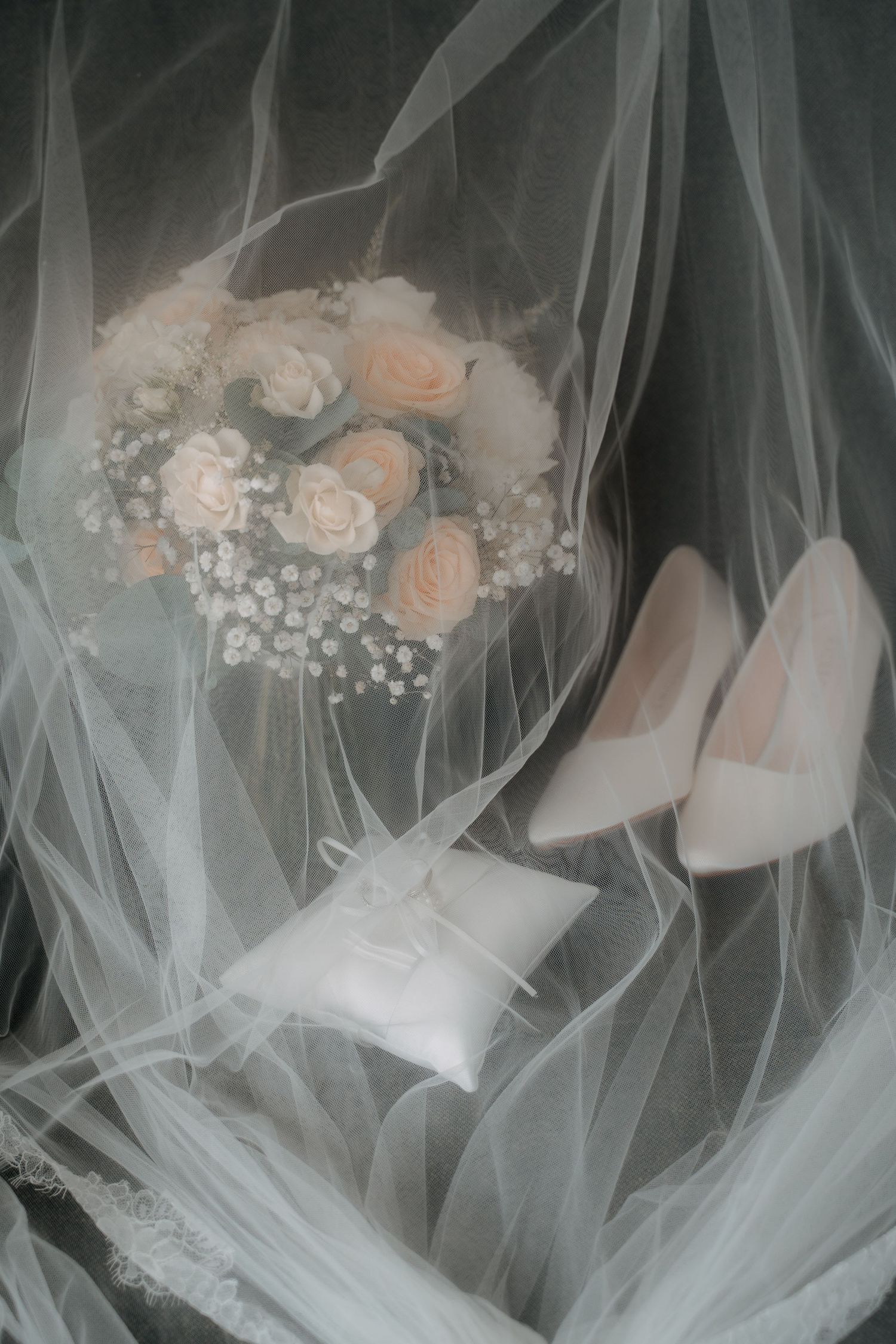 Bridal flat lay with white rose bouquet, ring pillow, and ivory heels arranged on flowing wedding veil.