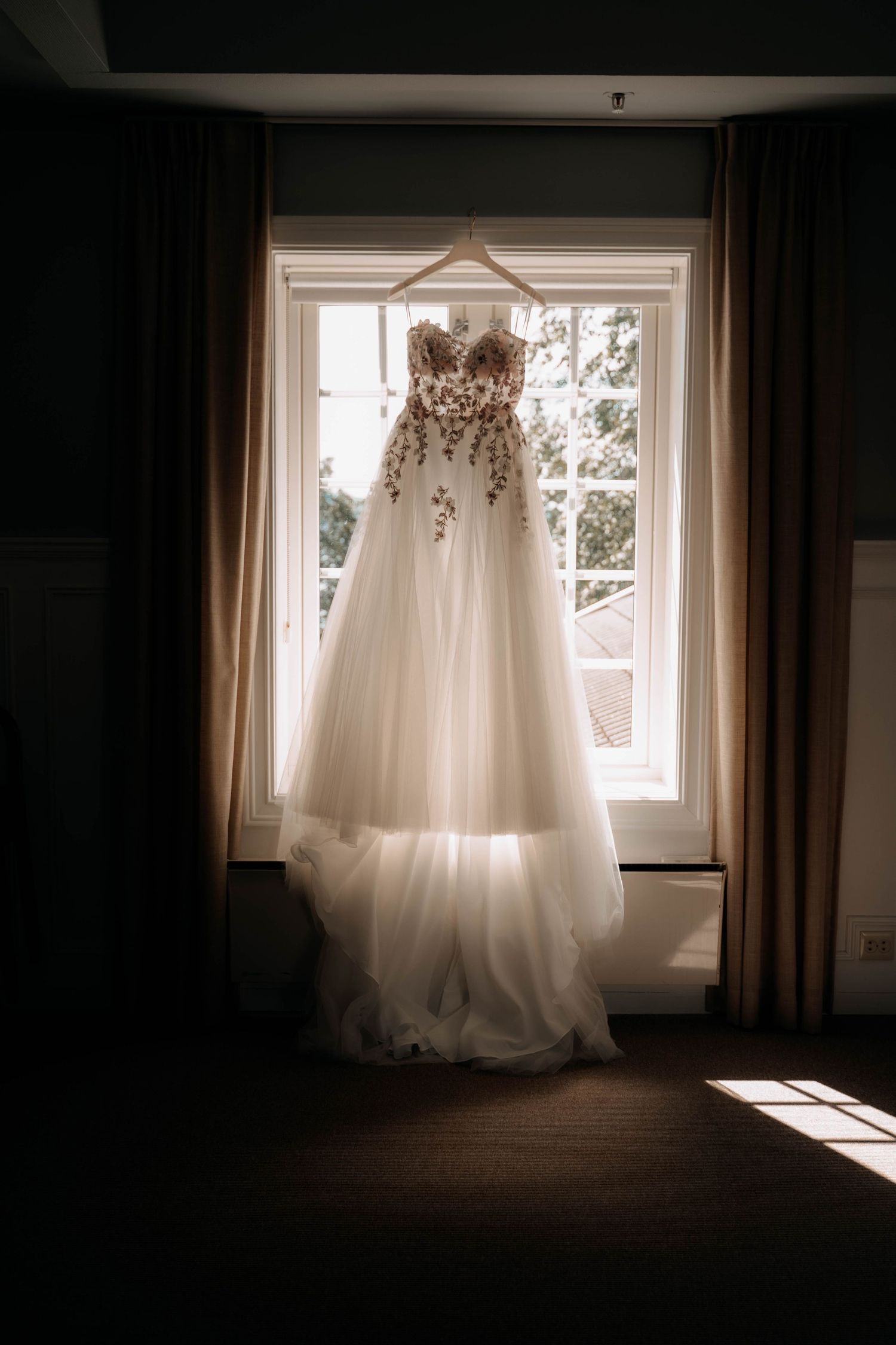 Elegant strapless wedding gown with floral embroidery hanging in a sunlit window before the ceremony.