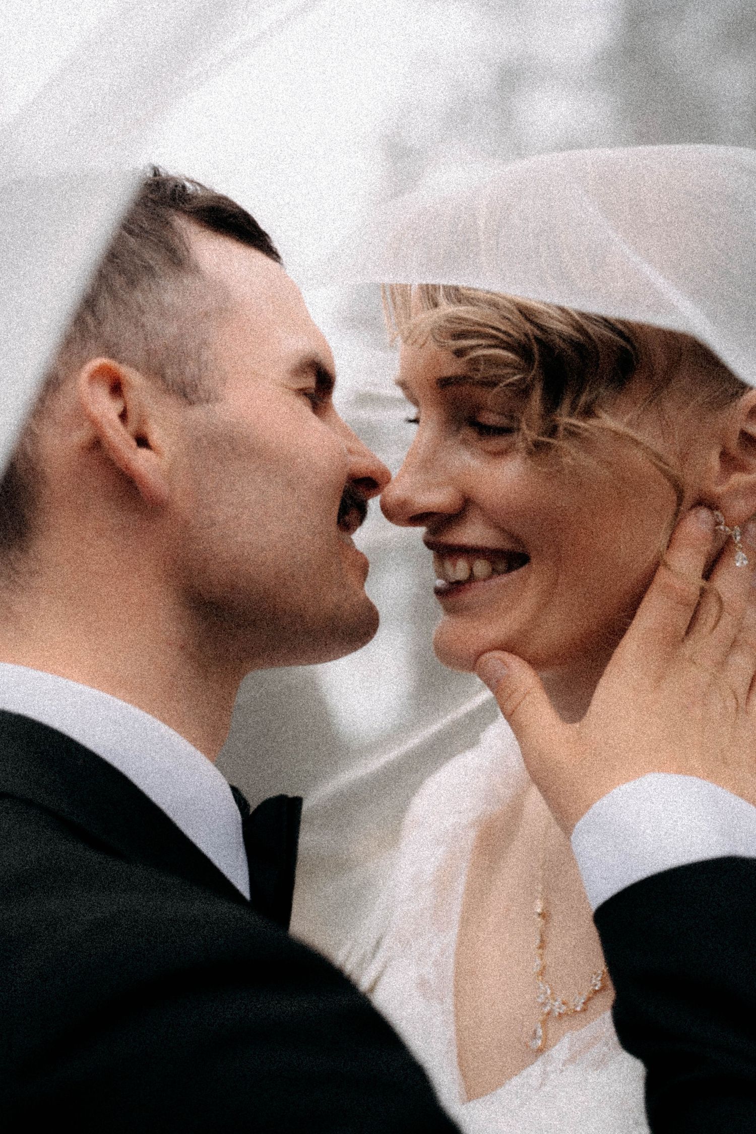 A bride and groom share an intimate moment under a veil, smiling close together on their wedding day.