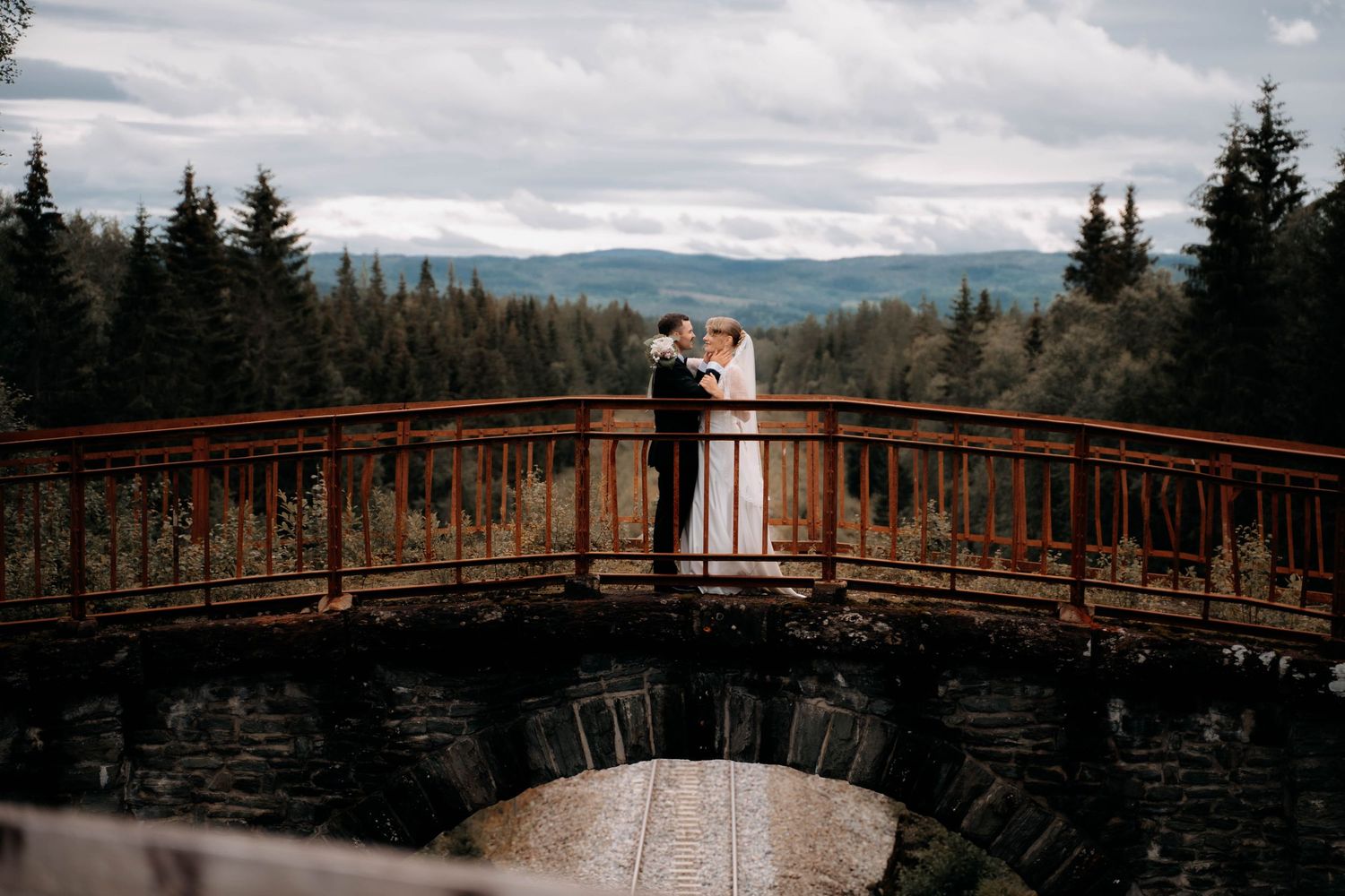 Wedding couple embracing on a rustic stone bridge surrounded by forest and mountain landscape.