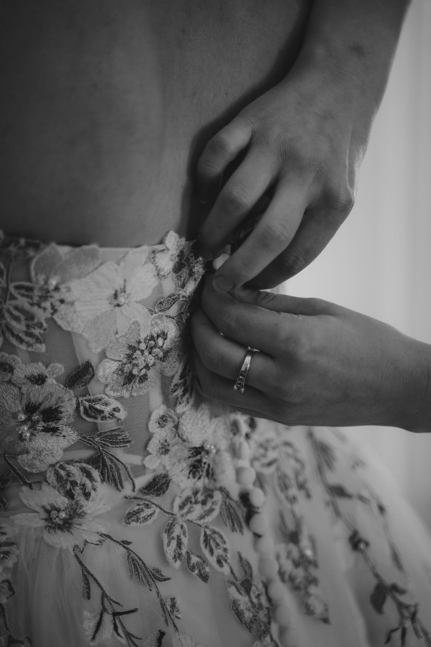 Black and white photo of hands fastening a floral lace wedding dress, with a diamond ring visible on one finger.