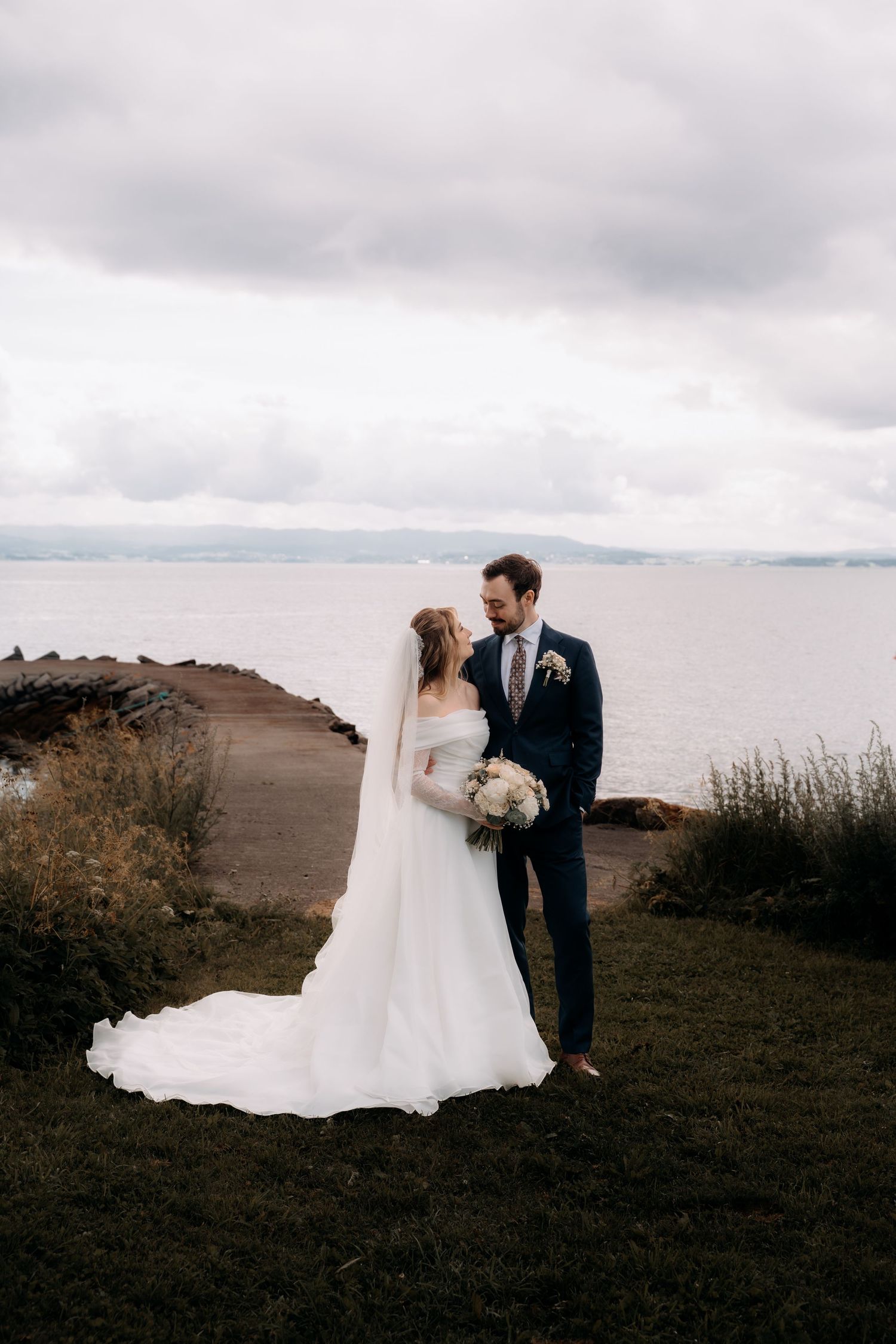 Bride and groom stand together by the waterfront, she in a white gown with veil, he in a navy suit holding her bouquet.