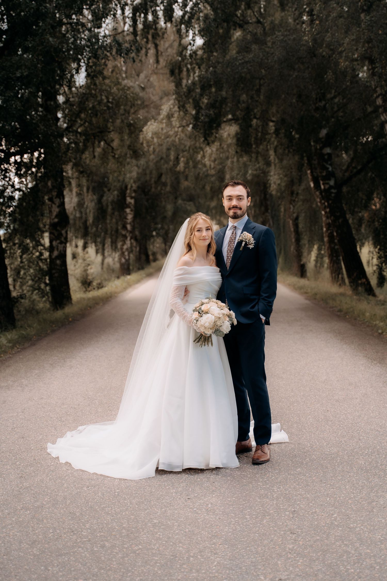 Bride in white off-shoulder gown with veil and groom in navy suit pose on tree-lined path holding bouquet.