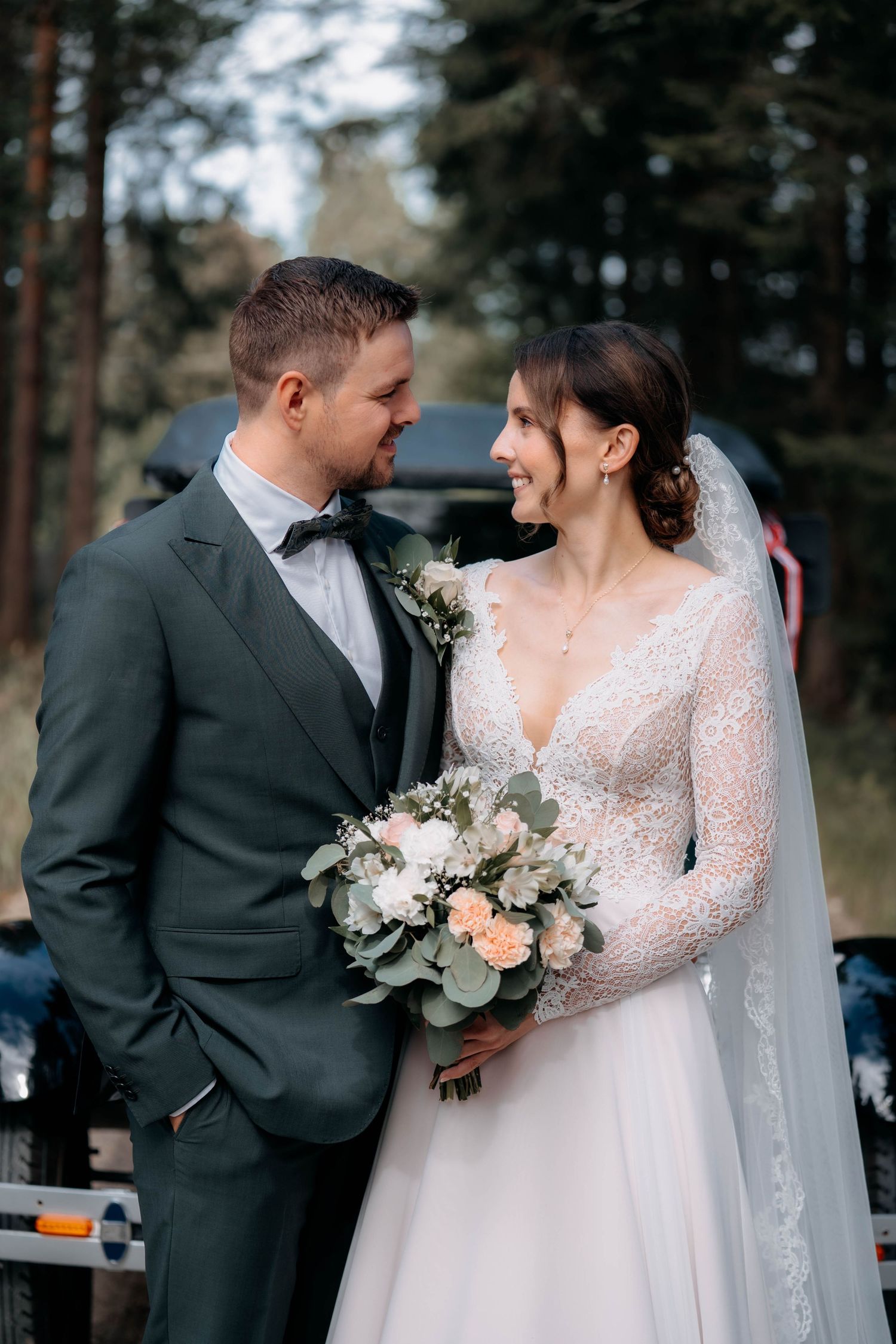 Bride and groom gazing at each other outdoors, she holds a bouquet of roses and eucalyptus, both dressed in wedding attire.