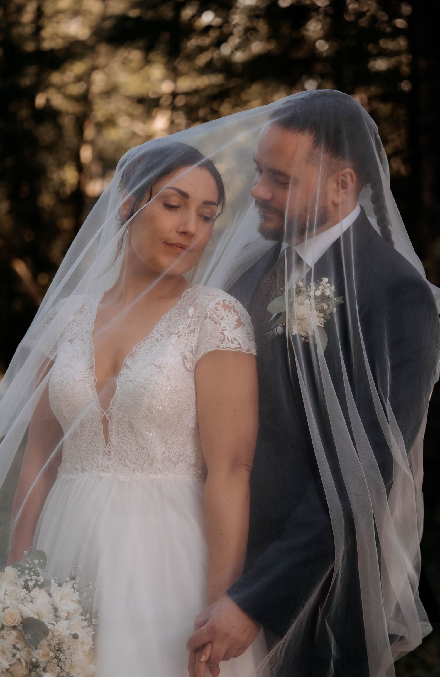 Bride and groom share a romantic moment under a flowing wedding veil, surrounded by a soft bokeh forest backdrop.