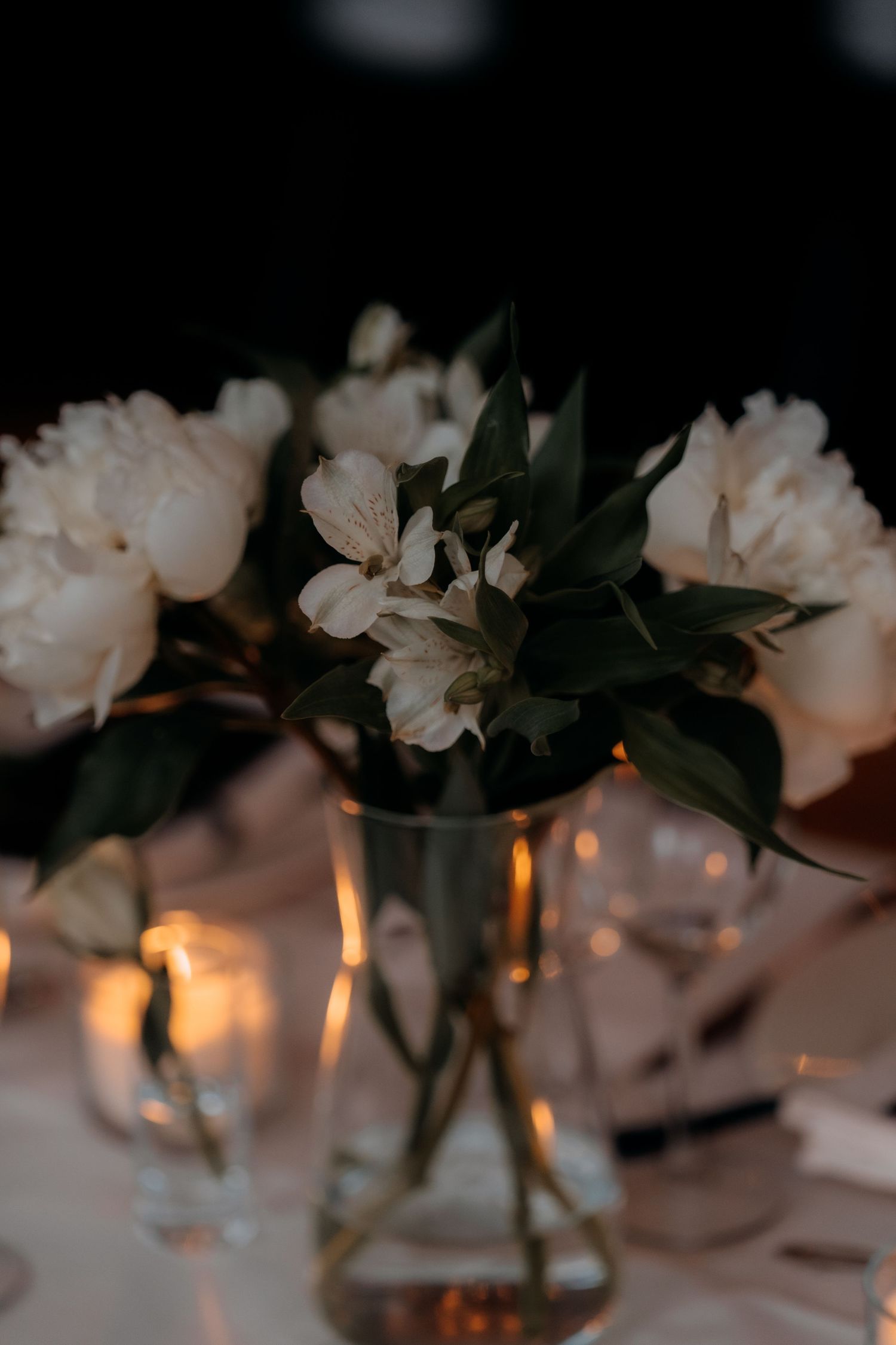 Elegant white peonies and alstroemeria in a glass vase centerpiece with candlelight on a reception table.