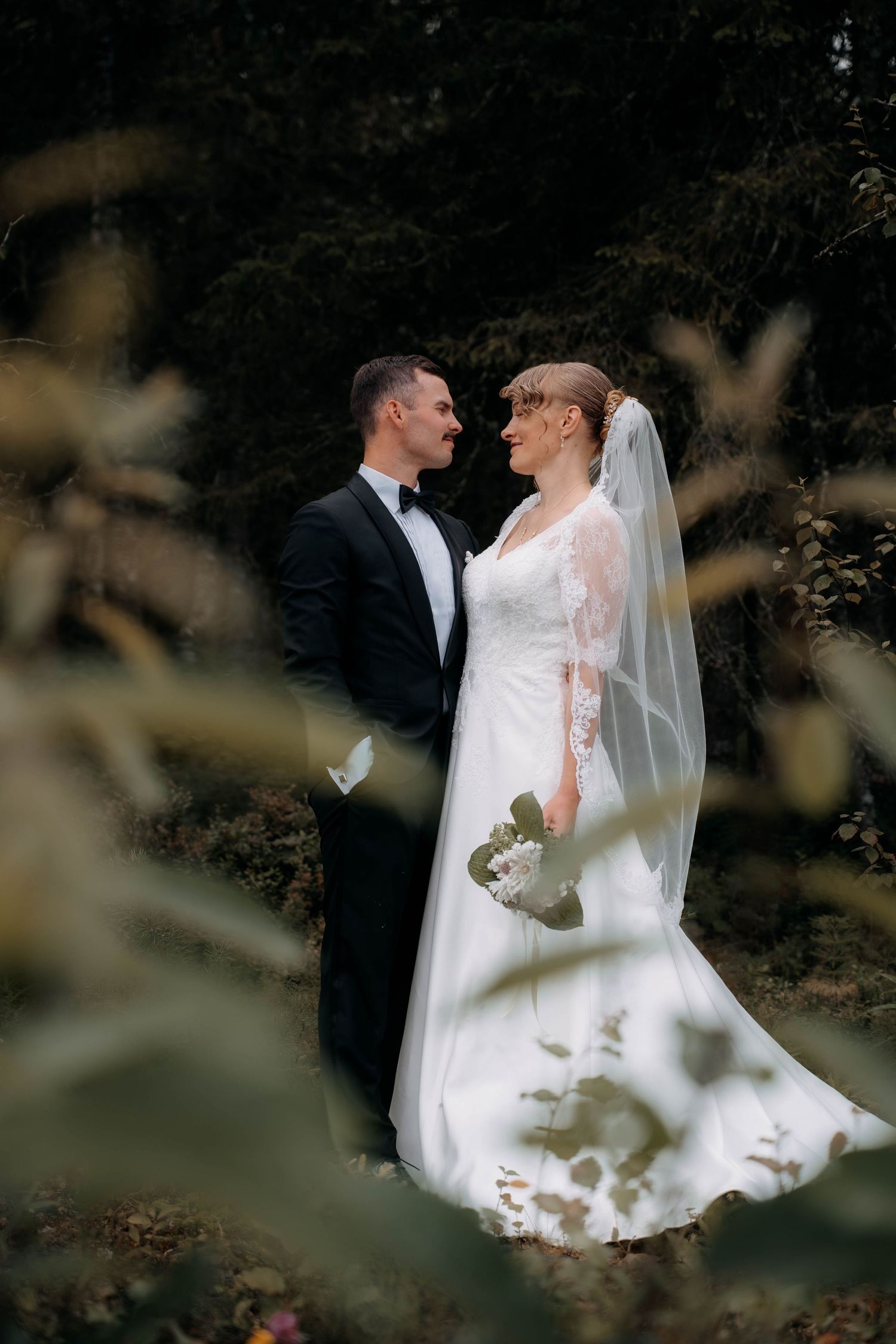 A bride and groom share a romantic moment in a lush forest setting, with the bride in a lace gown and veil.