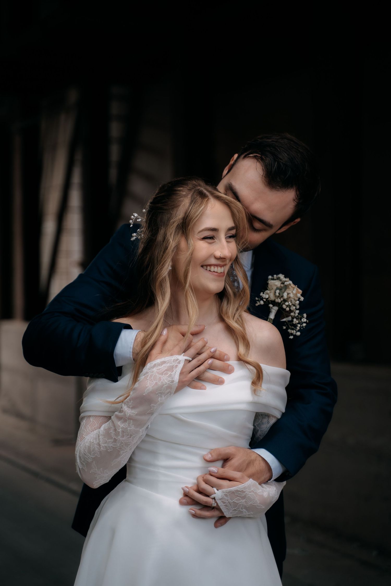 Groom in navy suit kissing bride in white off-shoulder lace gown, both smiling, elegant wedding portrait.
