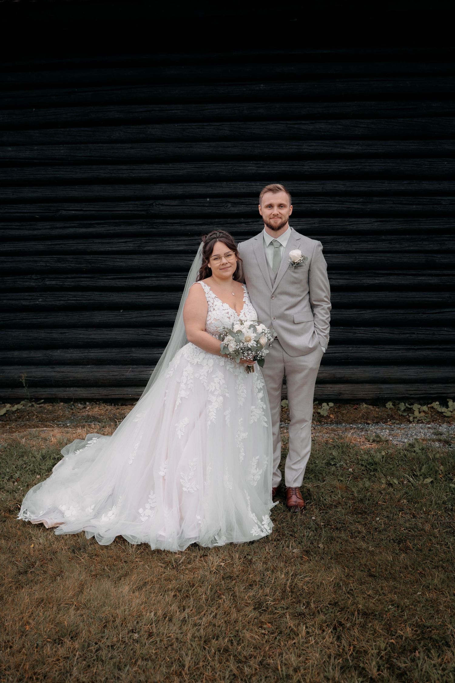 Bride in white lace gown with veil and groom in gray suit pose together outdoors before a dark barn wall.