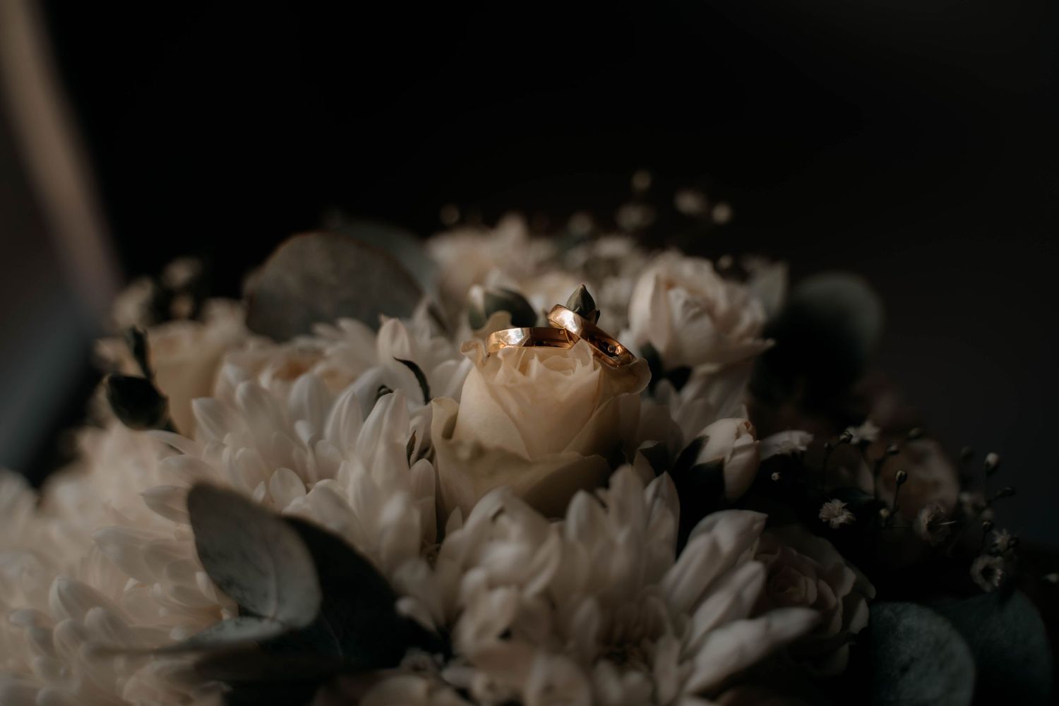 Two gold wedding rings rest on a bridal bouquet of white chrysanthemums, roses, and eucalyptus leaves.