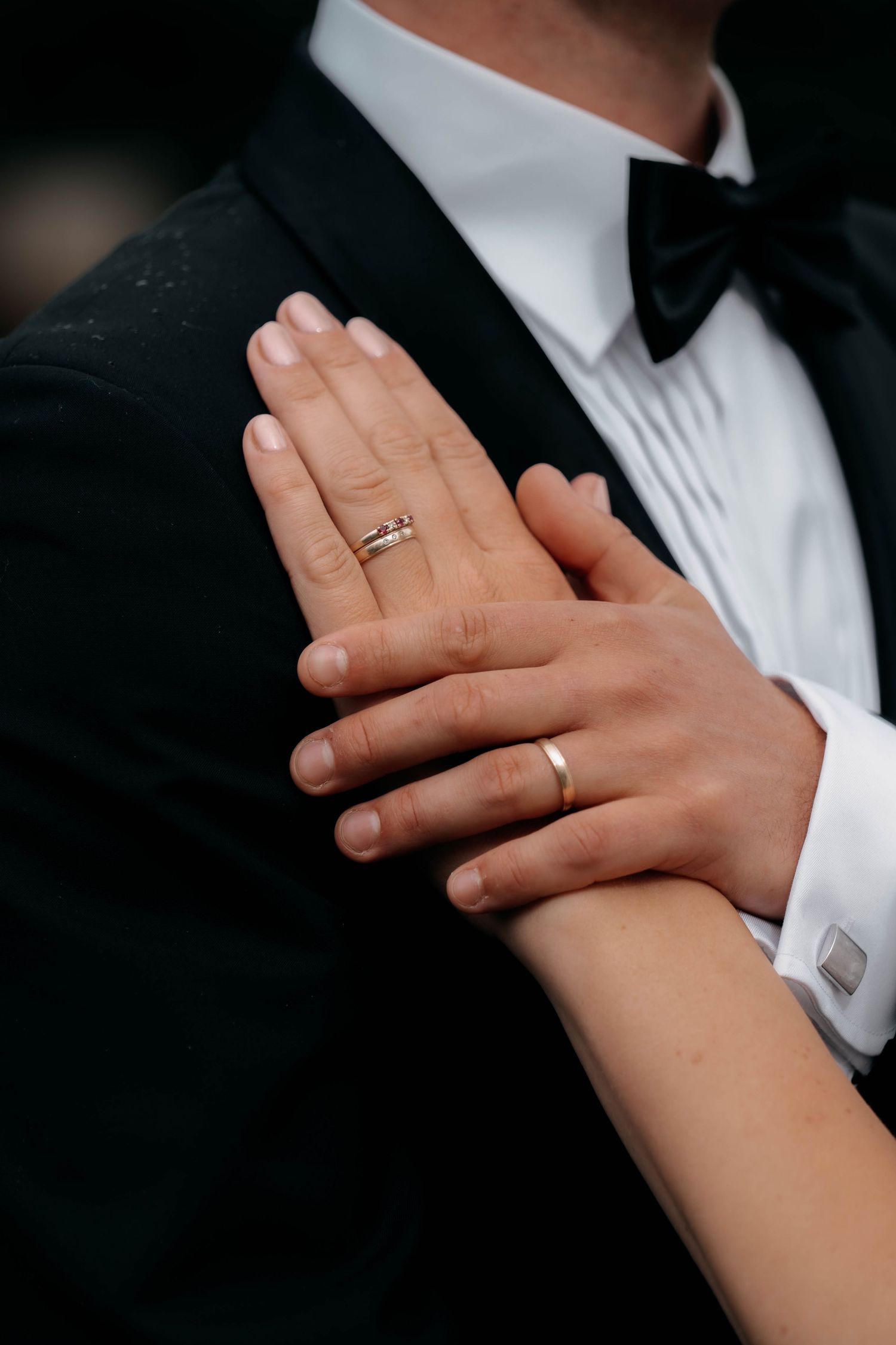 Close-up of newlyweds hands showing wedding rings, bride wearing rose gold band, groom in black tuxedo with white cufflinks.