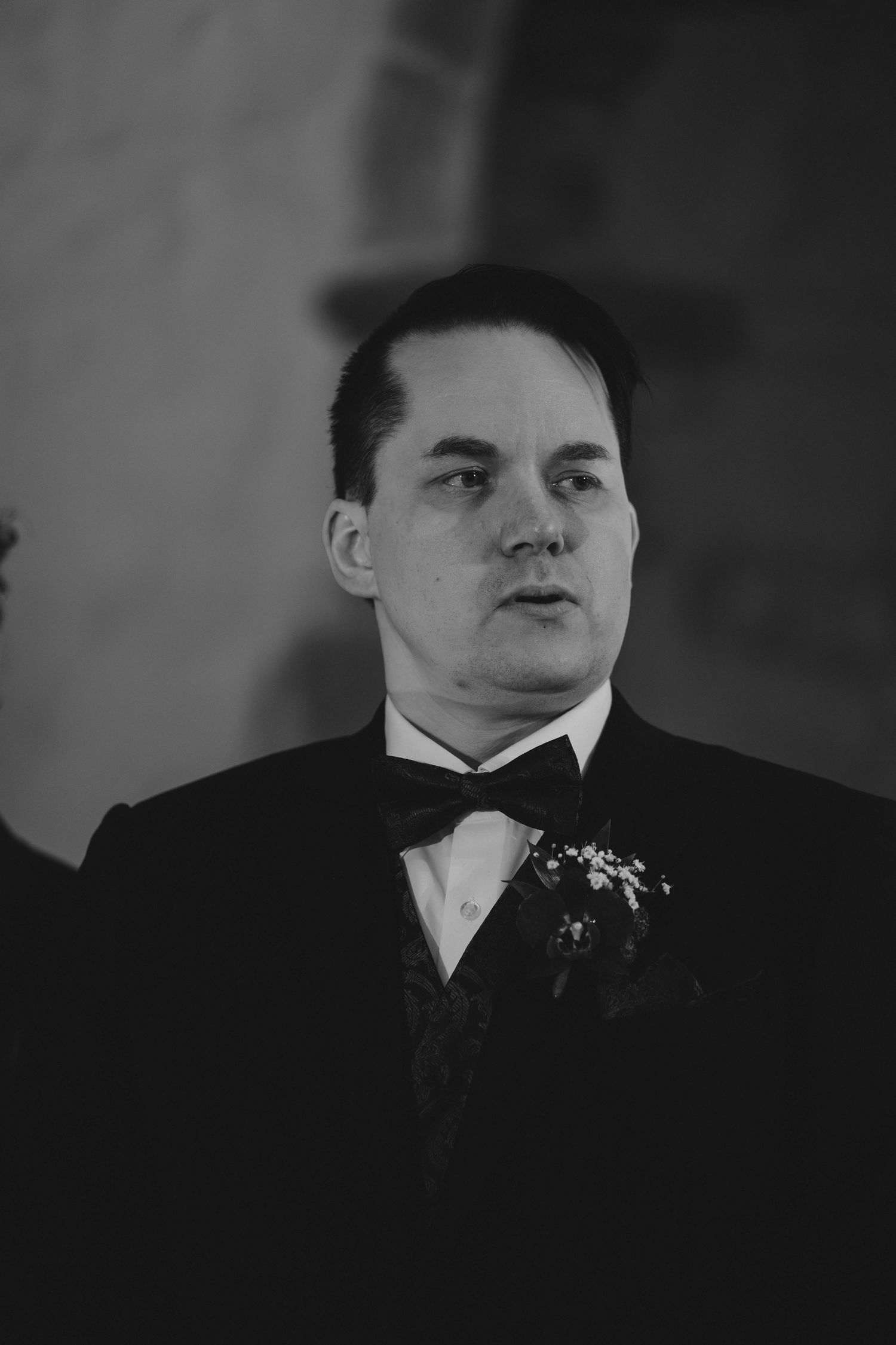 Black and white portrait of a groom in a tuxedo with boutonniere at a wedding ceremony.