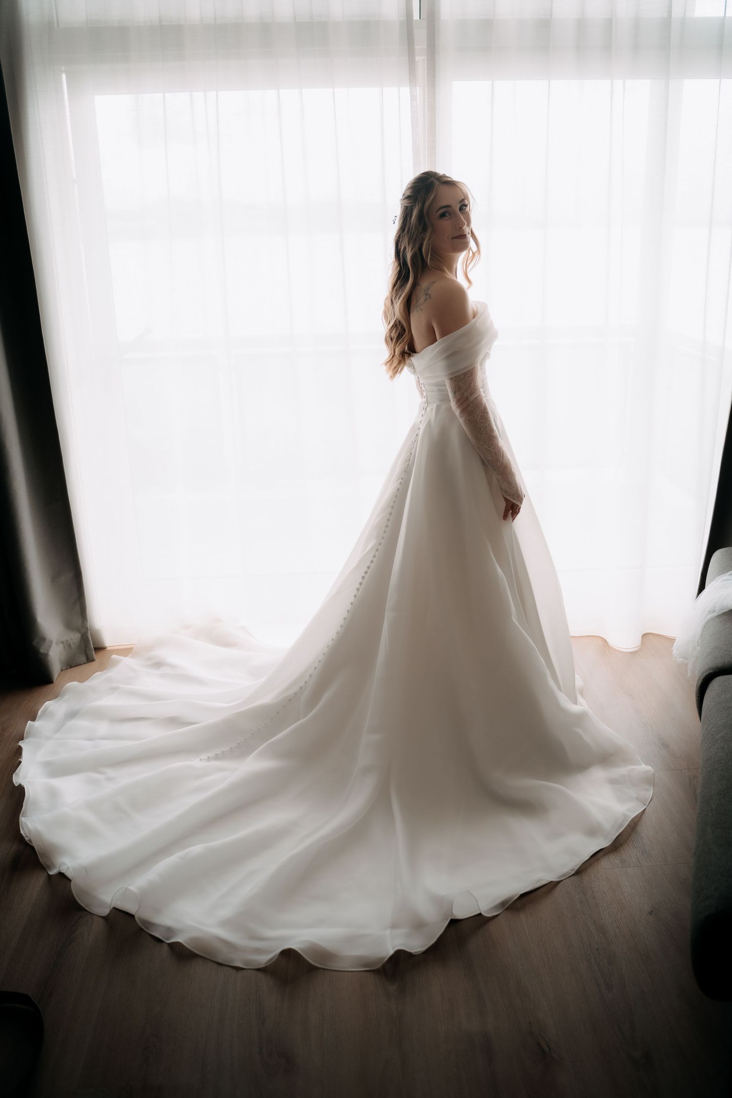 Bride in elegant off-shoulder white ball gown with long train standing by bright window on wedding day.