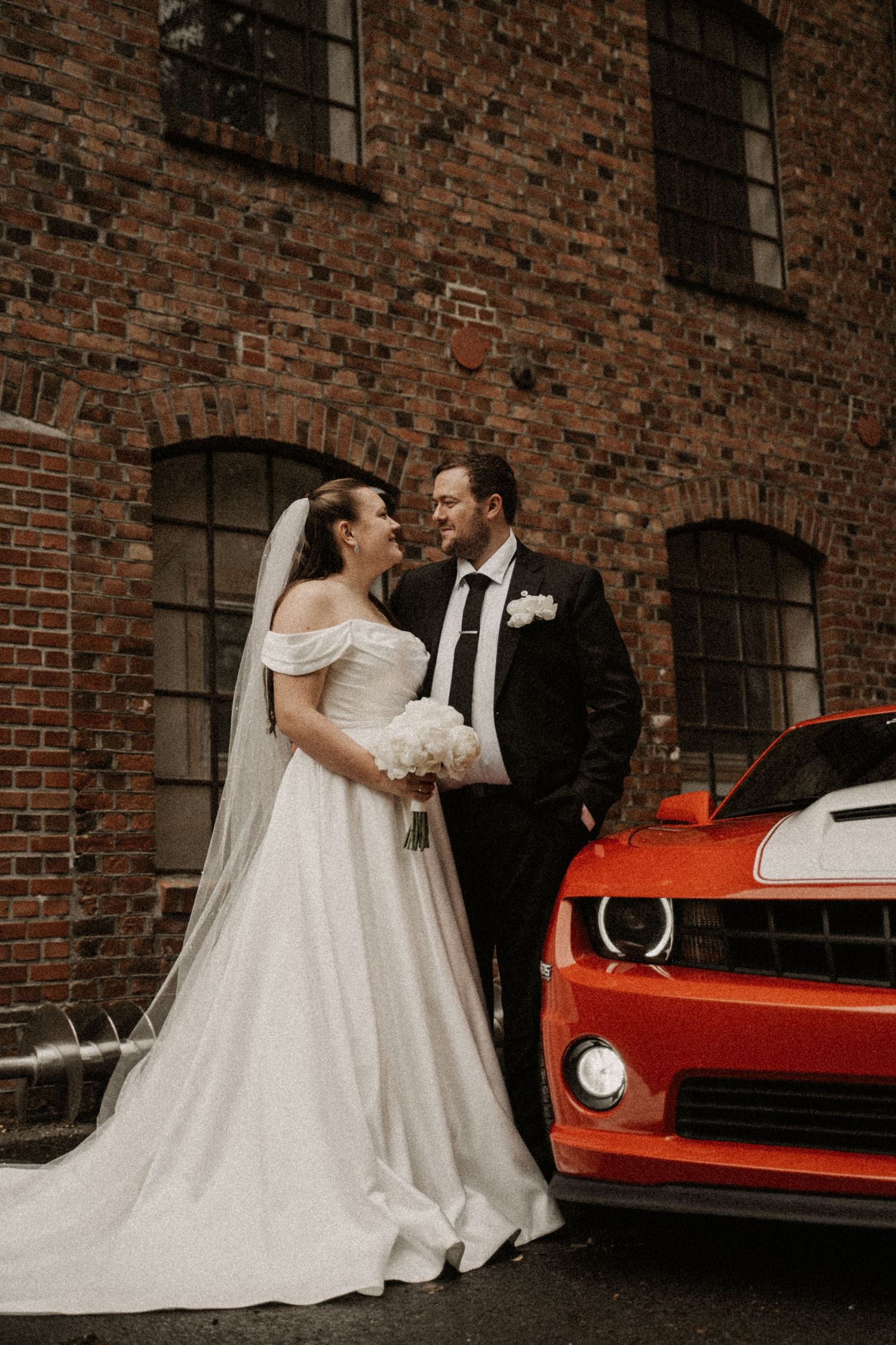 Bride and groom pose beside a vibrant orange Camaro against a rustic brick building on their wedding day.