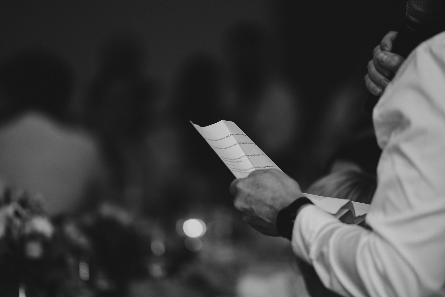 Man in white dress shirt reading notes from paper while giving a speech at a formal event.