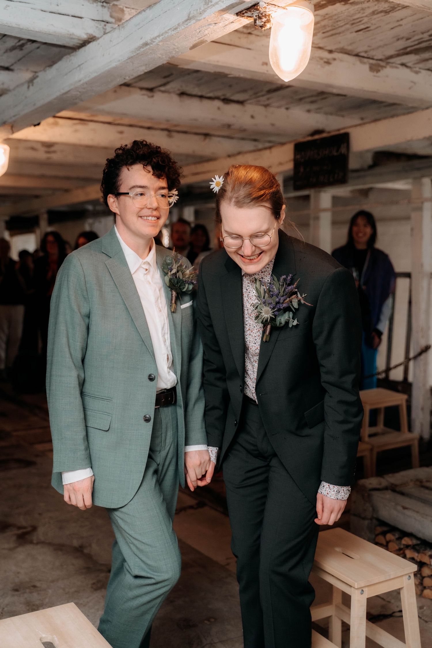 Two brides in suits walking down the aisle, smiling, wearing boutonnieres at their wedding ceremony.