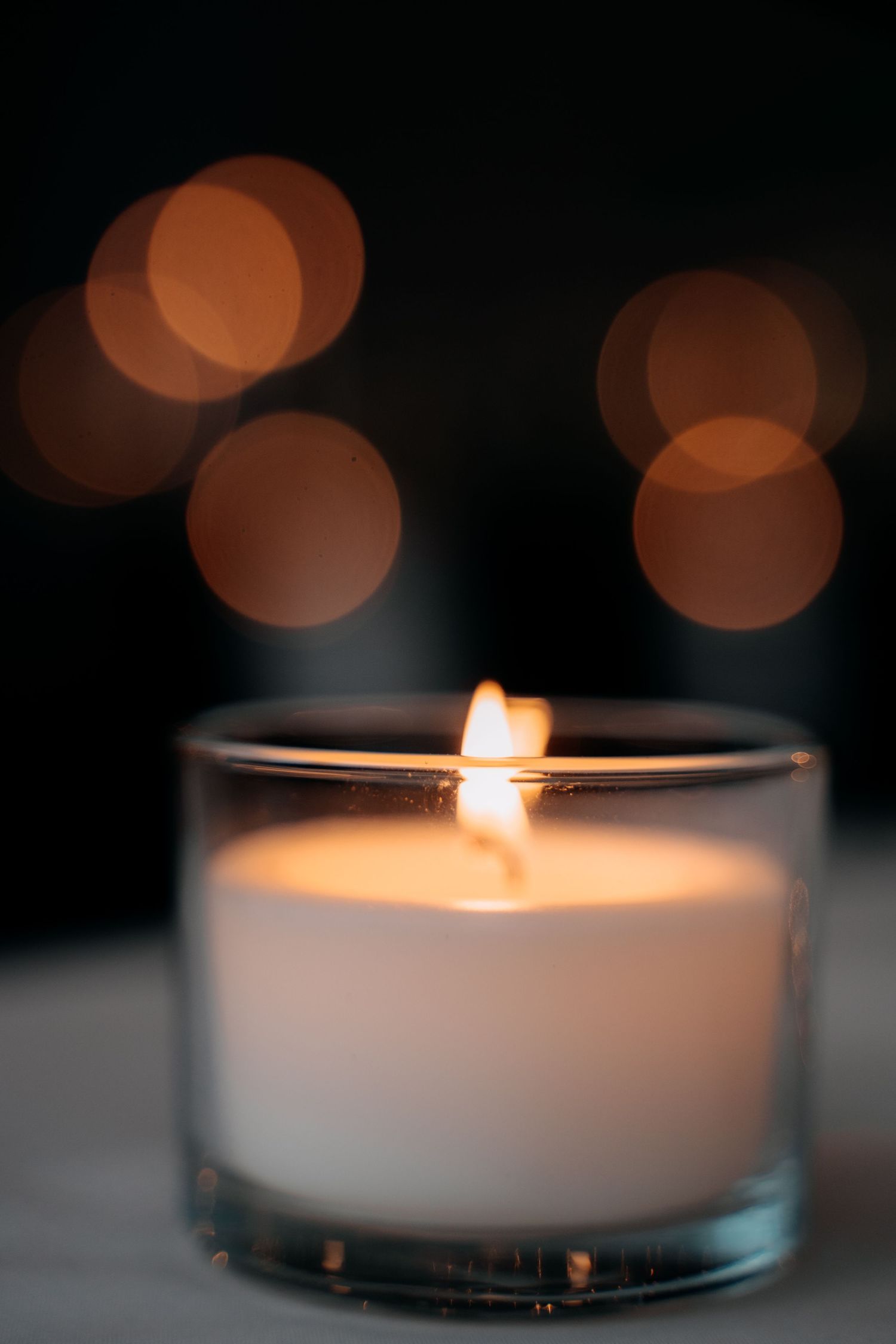 White candle in glass holder glowing warmly with golden bokeh lights in dark background for romantic ambiance.