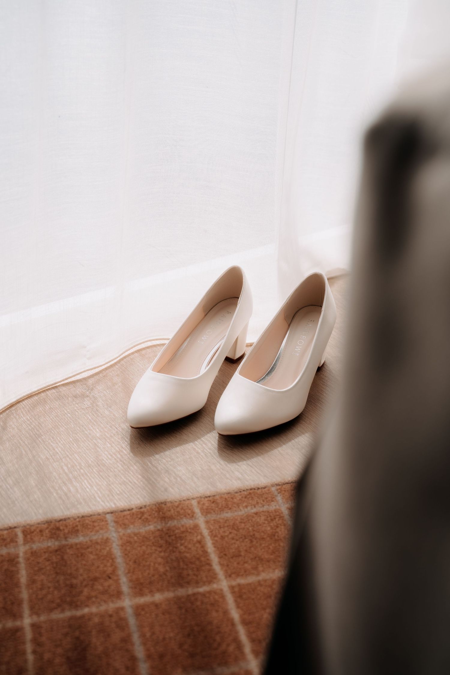 Elegant white block-heel bridal shoes placed on a brown plaid rug near a white curtain in a softly lit room.
