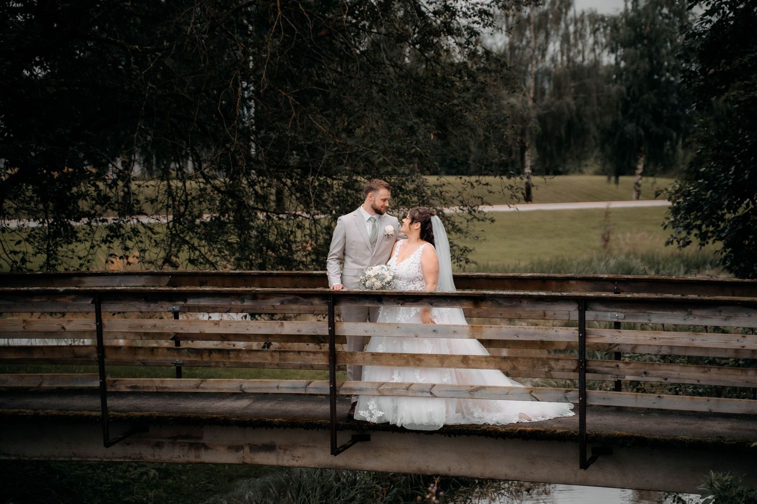 Bride and groom share a romantic moment on a wooden bridge surrounded by lush green trees on their wedding day.