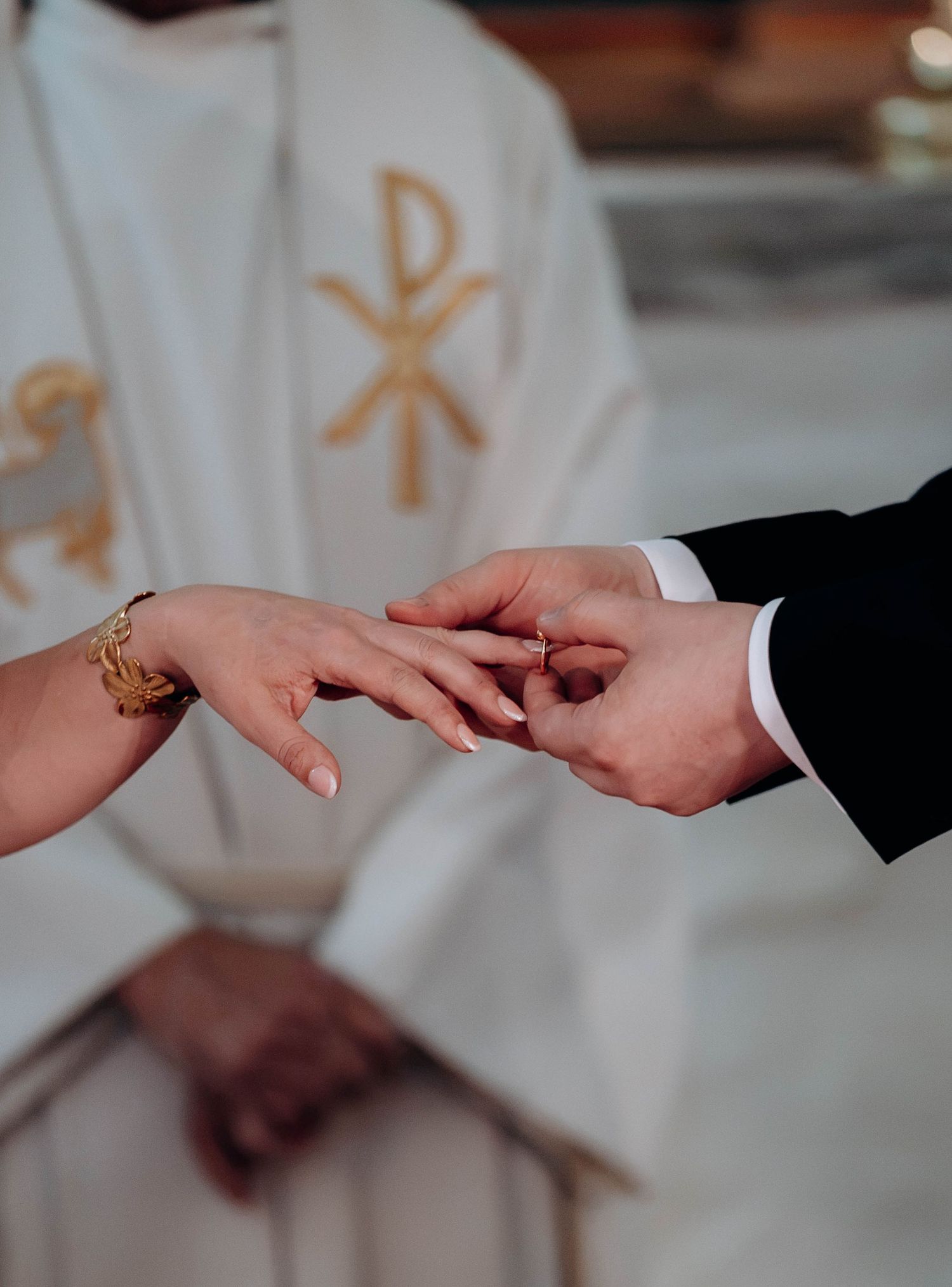 Groom placing a wedding ring on bride's finger during a church ceremony with priest in white vestments.