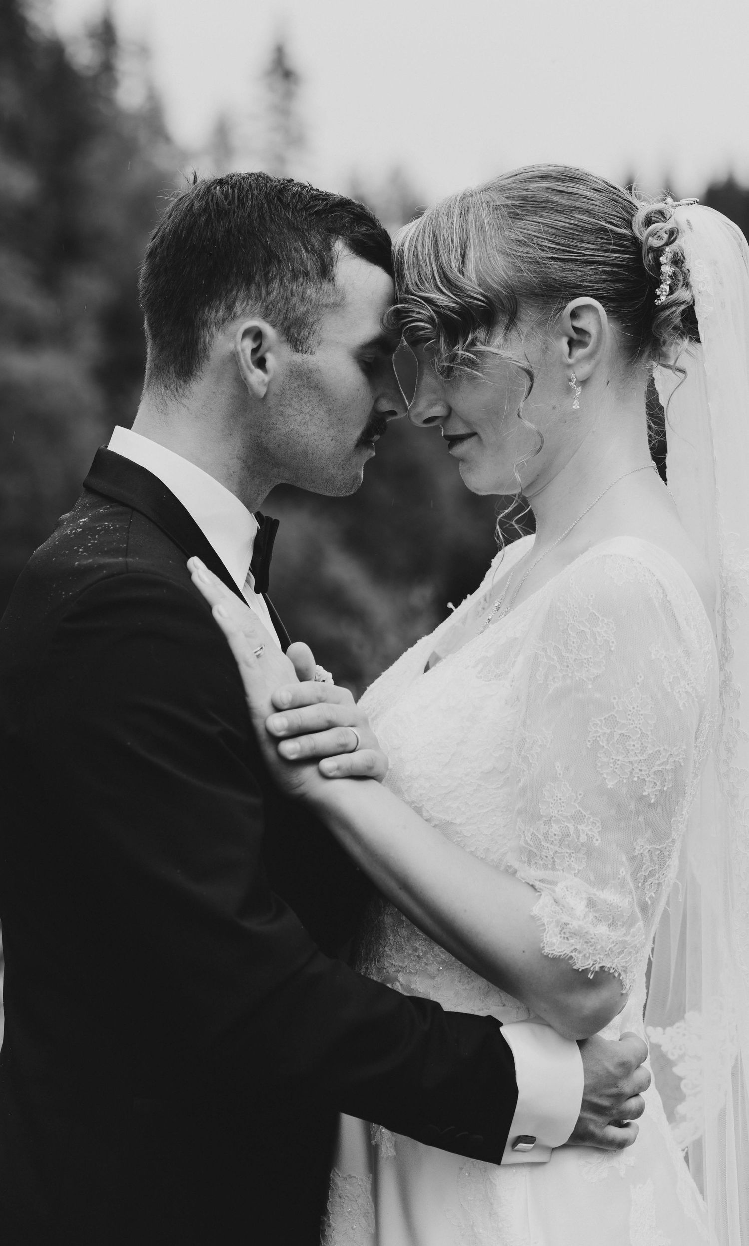 Black and white romantic portrait of bride and groom in tender embrace, foreheads touching, surrounded by pine trees.