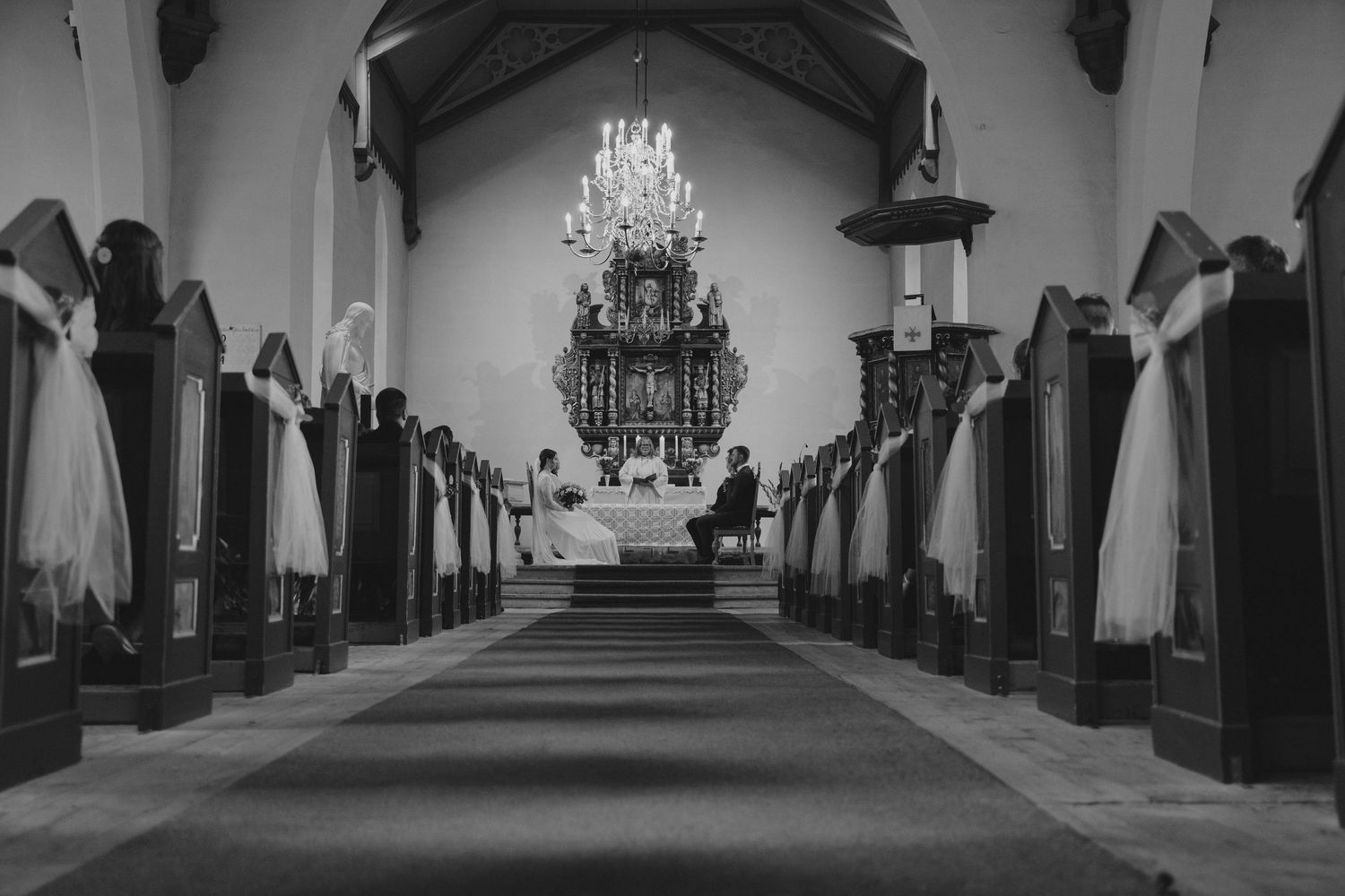 Black and white photo of elegant church wedding ceremony with chandelier and decorated pews.