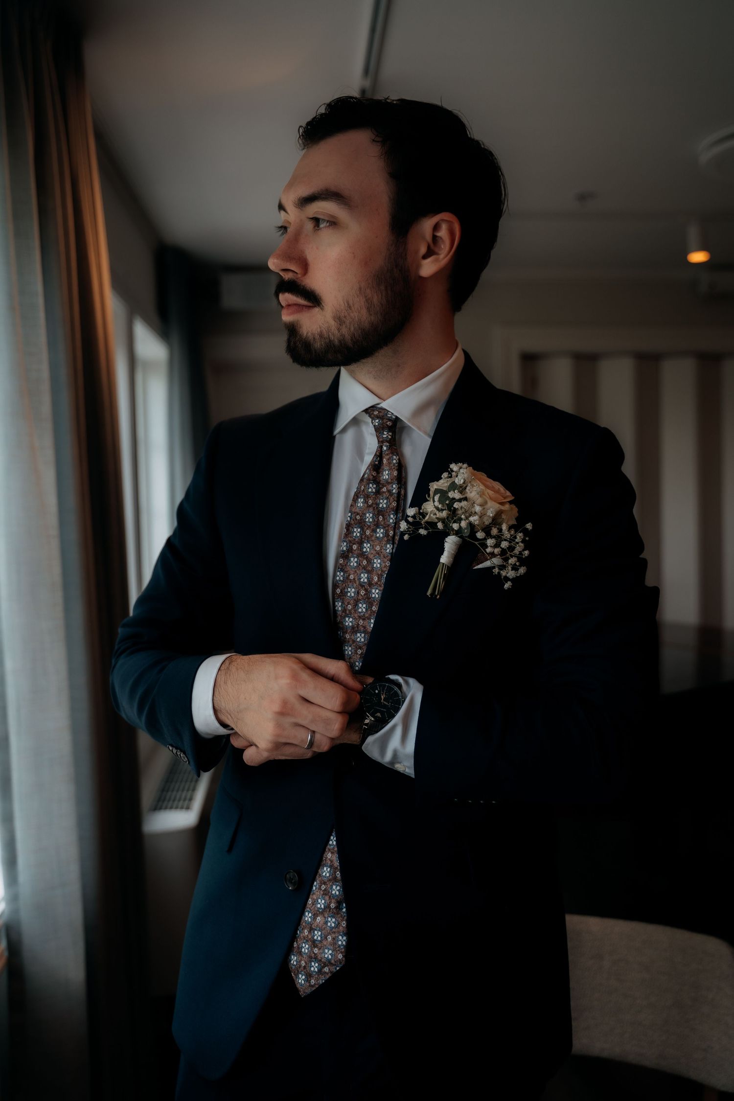 Groom in navy suit adjusting watch, wearing floral boutonniere and patterned tie, standing by window on wedding day.