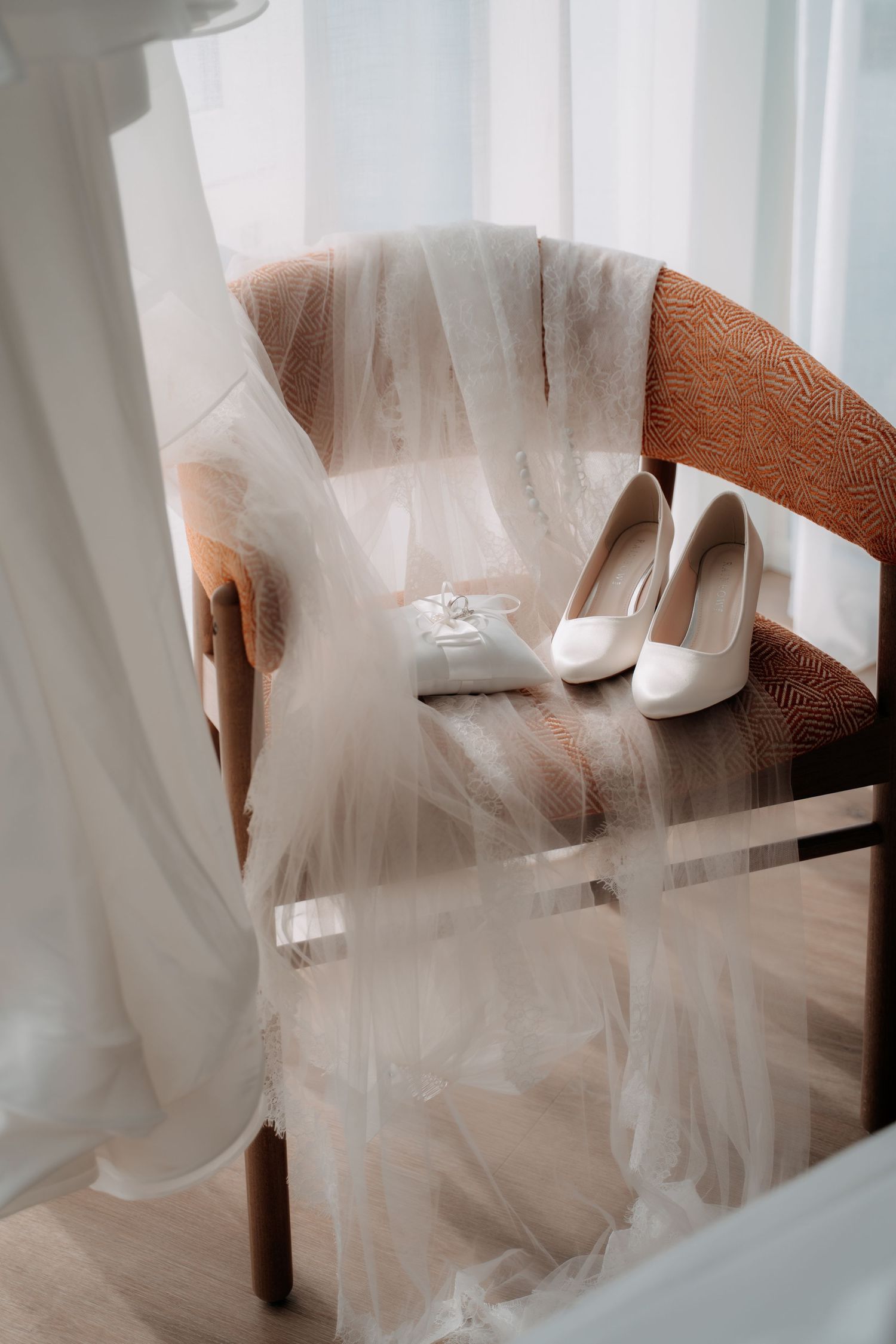 White bridal heels and ring box rest on a brown chair draped with a sheer wedding veil near a window.