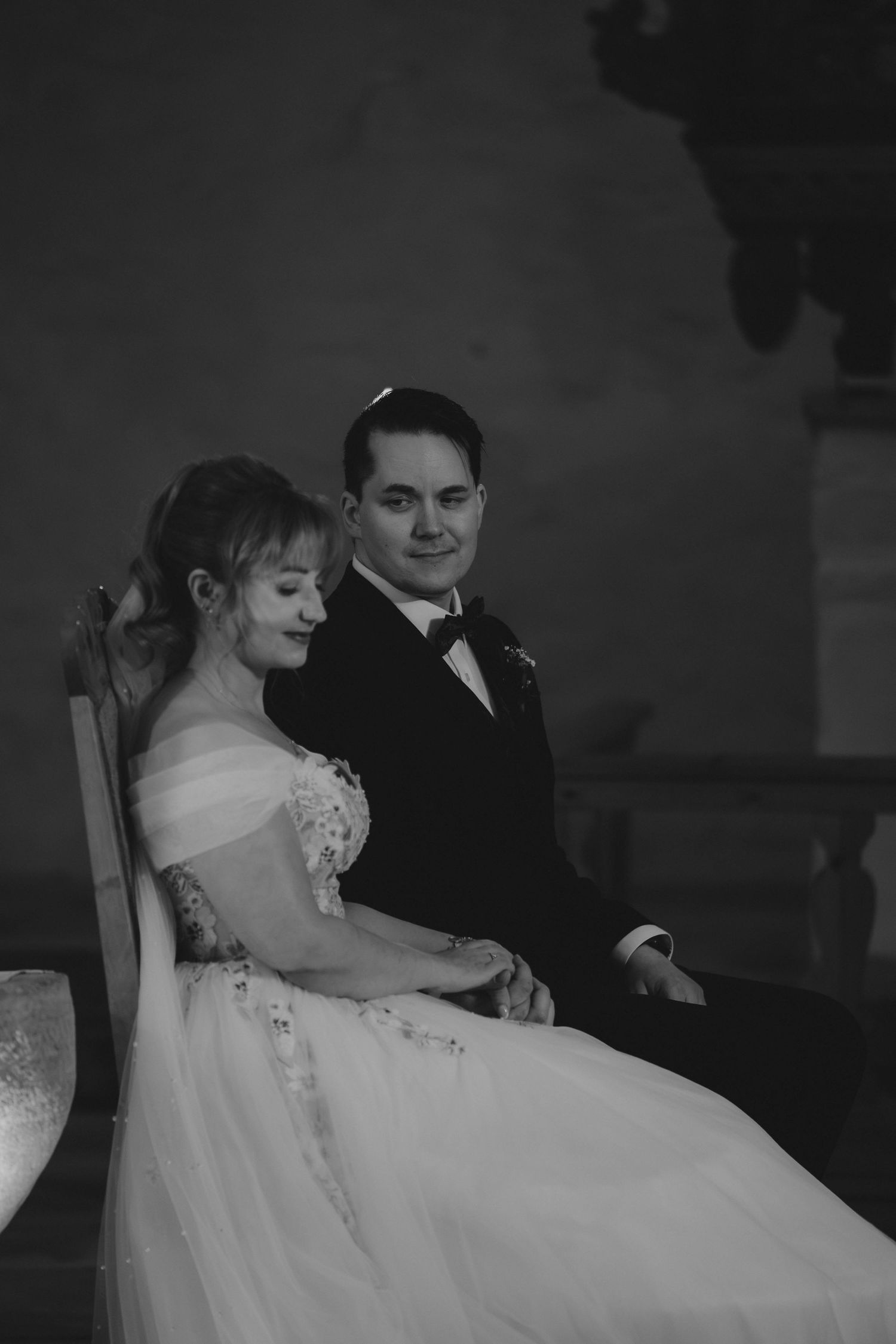 Black and white wedding photo of bride in off-shoulder gown and groom in tuxedo sitting together in elegant venue.