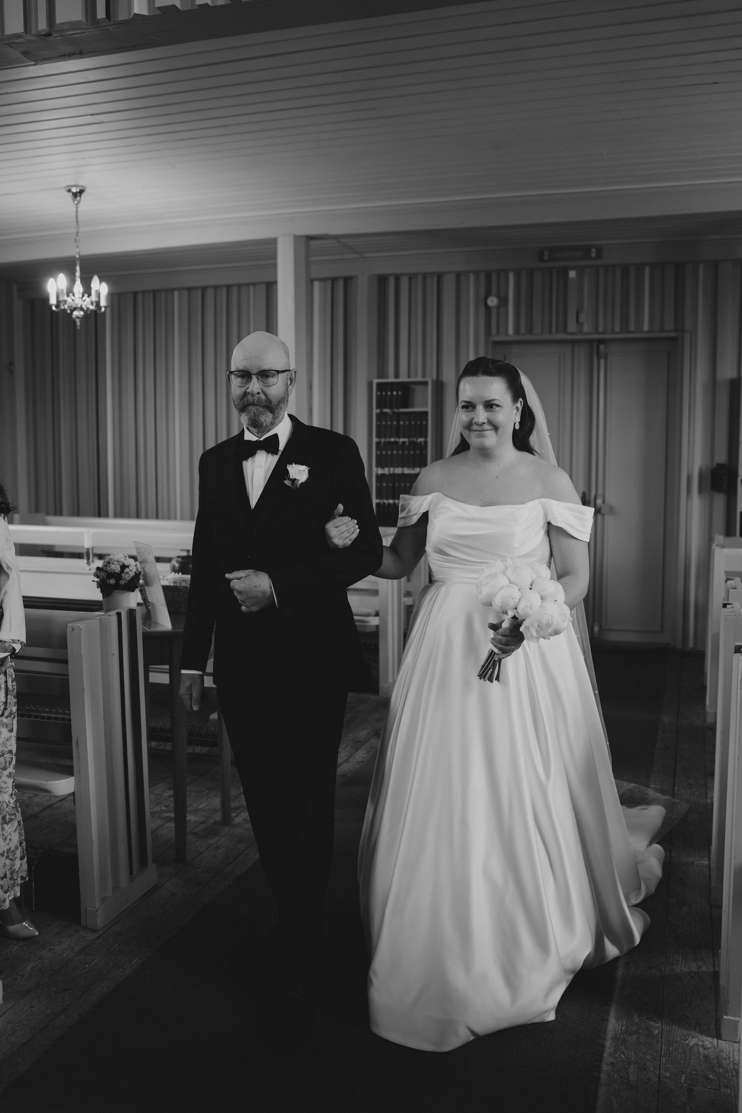 Black and white photo of a bride in an off-shoulder gown walking down the aisle with her father in a church.