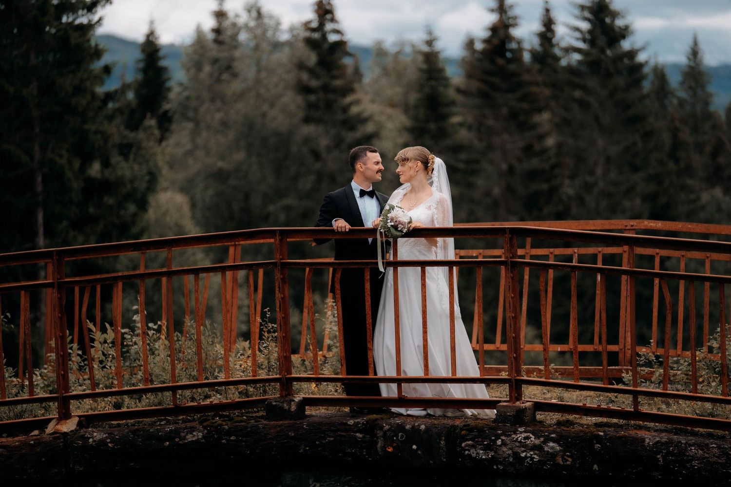 Bride and groom stand on a rustic red bridge surrounded by lush pine forest, sharing a romantic moment on their wedding day.