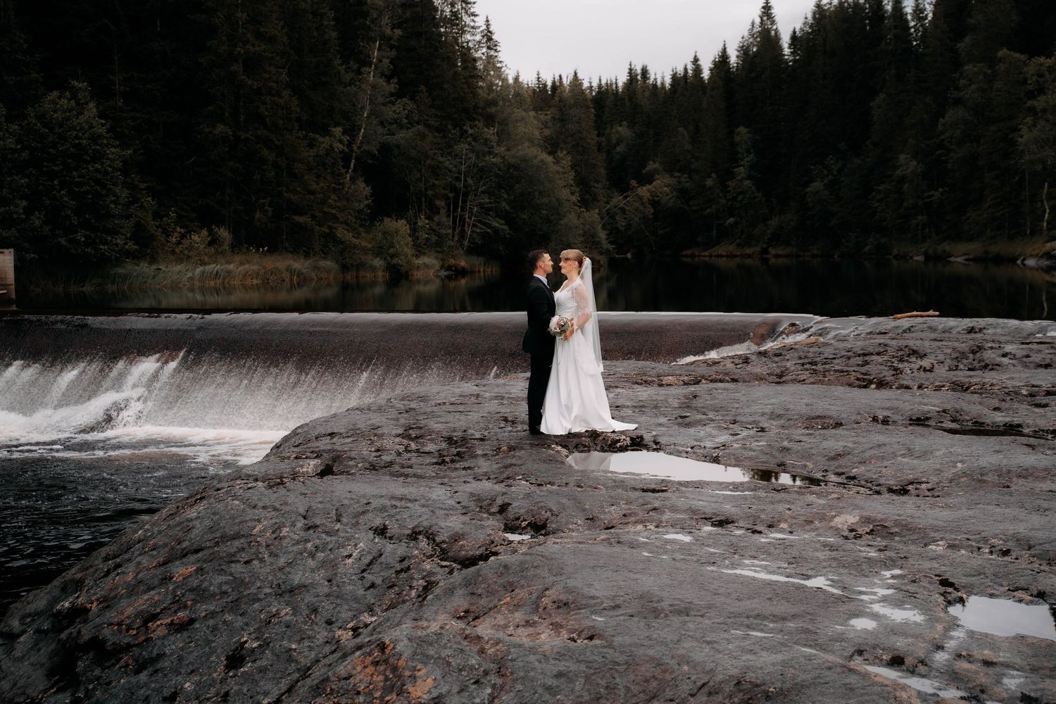 A bride and groom embrace on rocky terrain beside a cascading waterfall surrounded by lush evergreen forest.