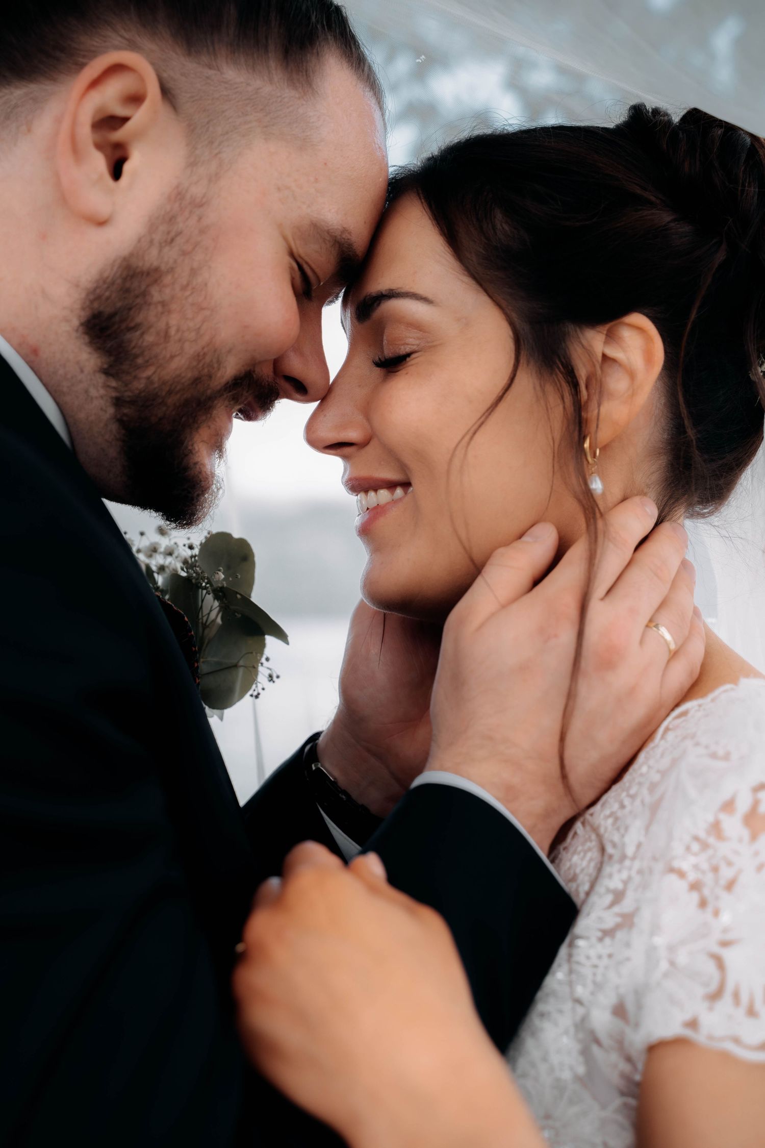 Romantic wedding couple touching foreheads, smiling bride in lace dress with pearl earring, groom in suit with boutonniere.