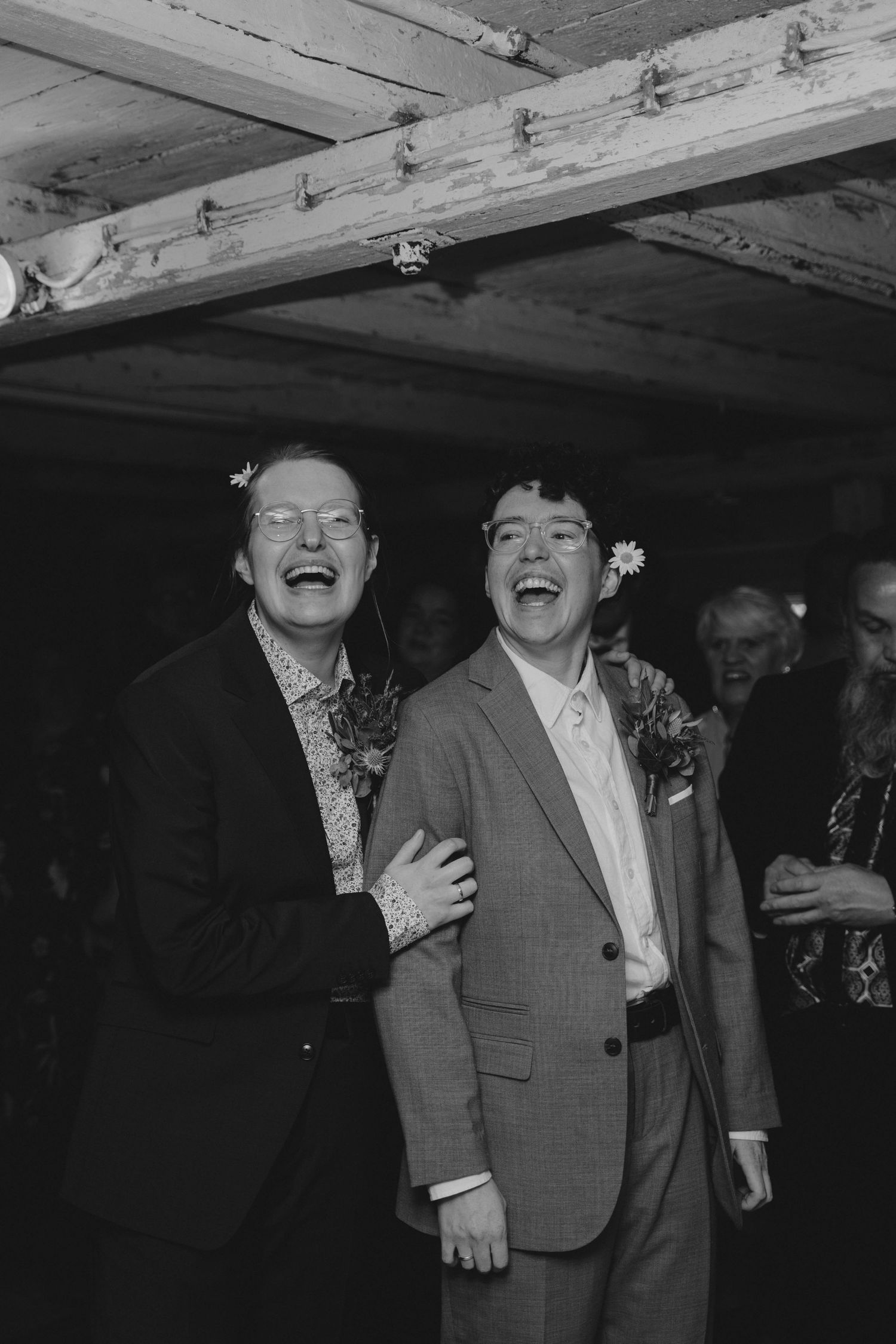 Two men laughing joyfully at a wedding reception, wearing suits with floral boutonnieres, in a black and white photo.