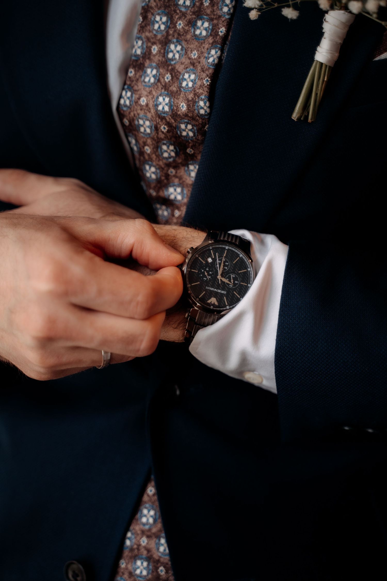 Man in navy suit adjusting black chronograph watch, wearing patterned tie and floral boutonniere.