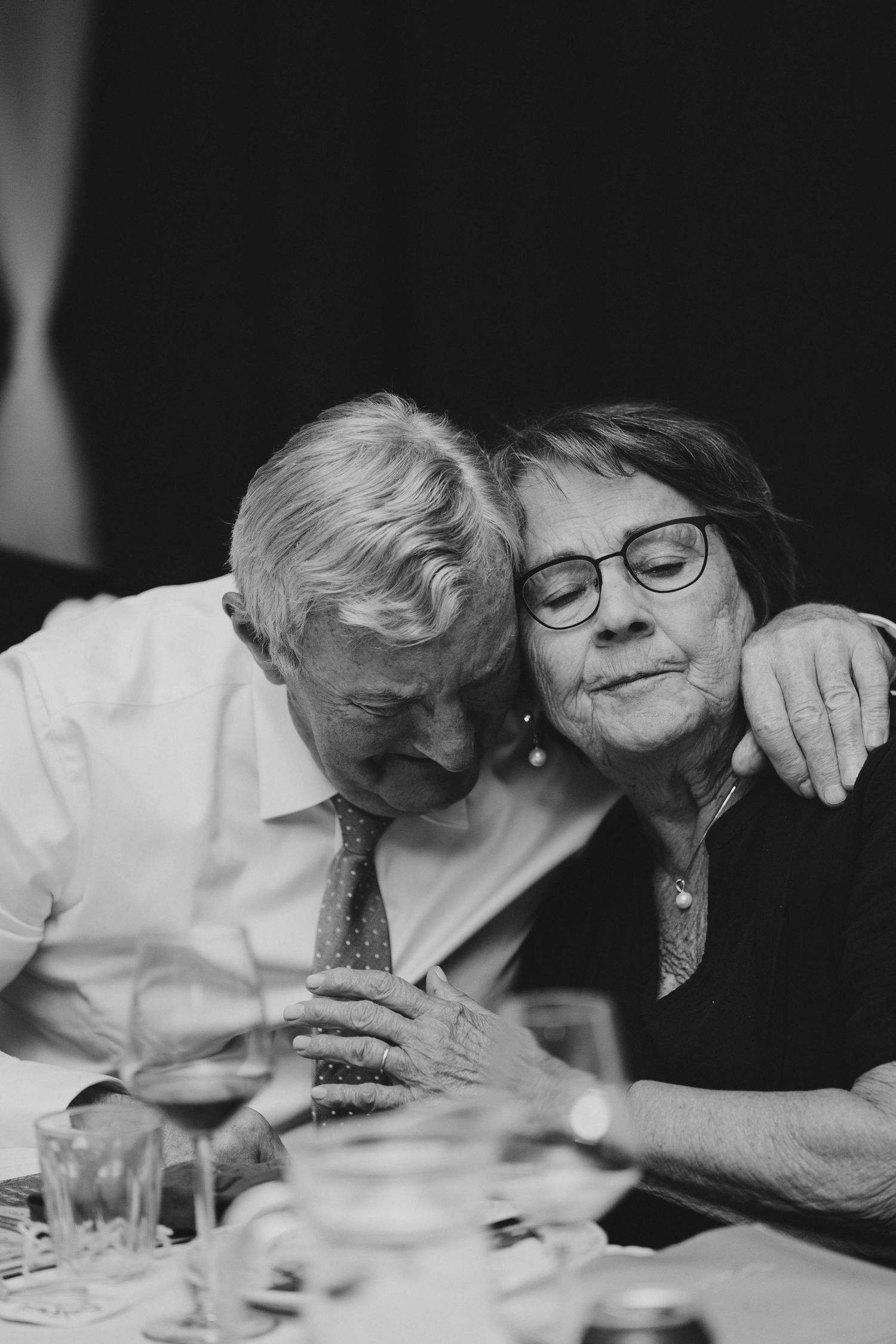 Elderly couple sharing a tender moment at a dinner table, leaning heads together in black and white photo.