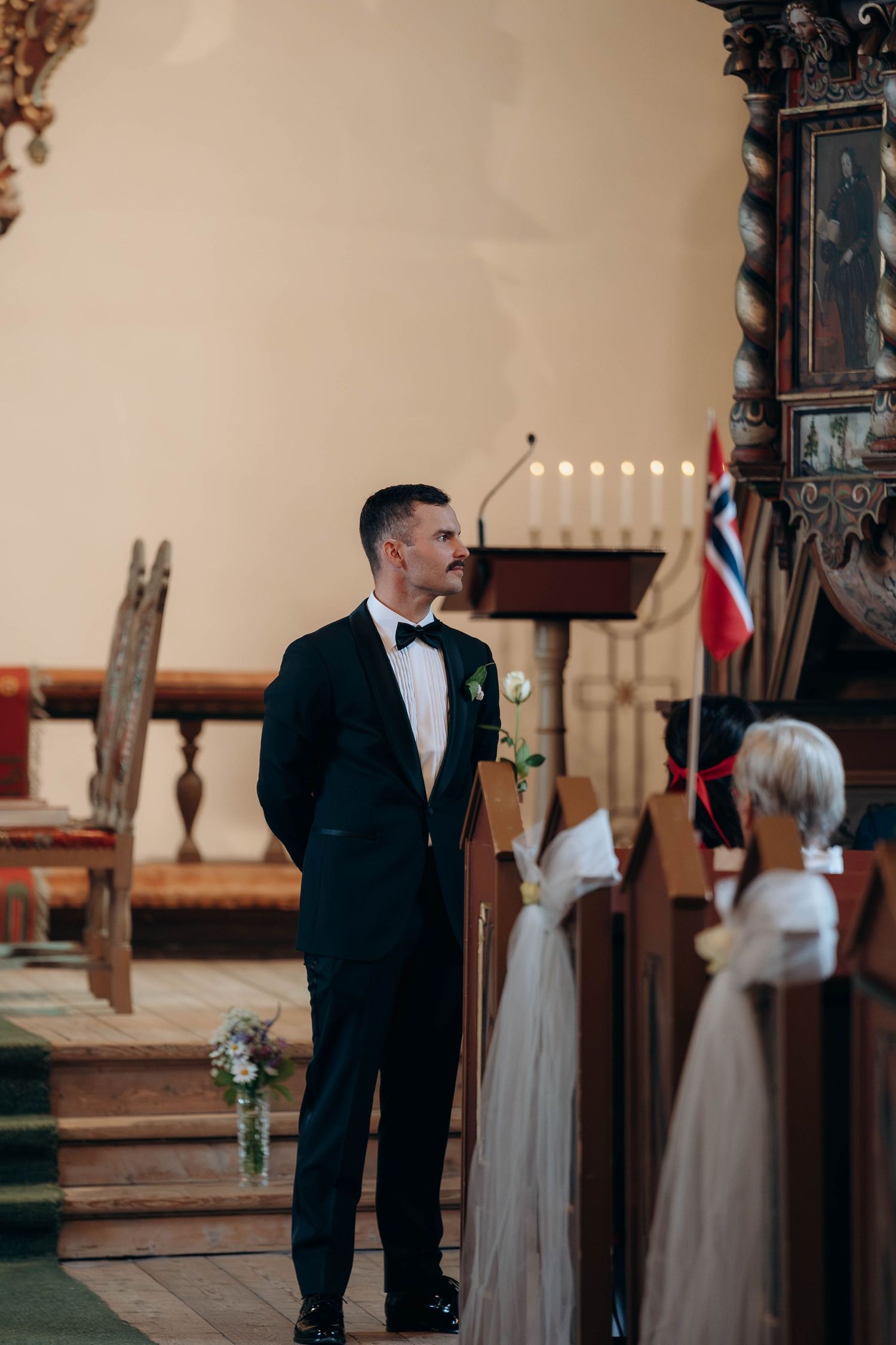 Groom in black tuxedo standing at church altar awaiting bride, Norwegian flag visible in background.