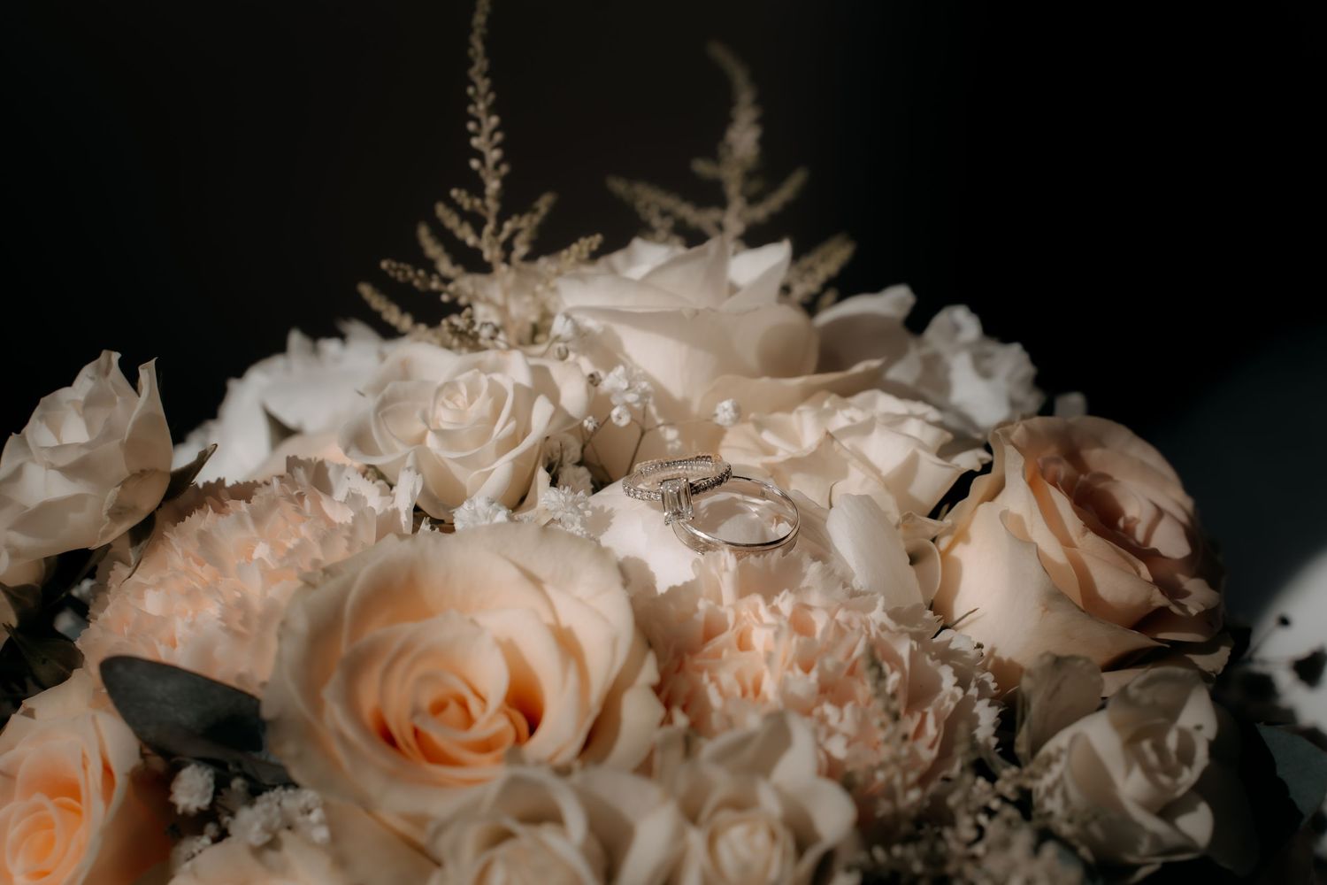 Wedding rings resting on a bridal bouquet of soft peach and white roses with delicate silver accents.