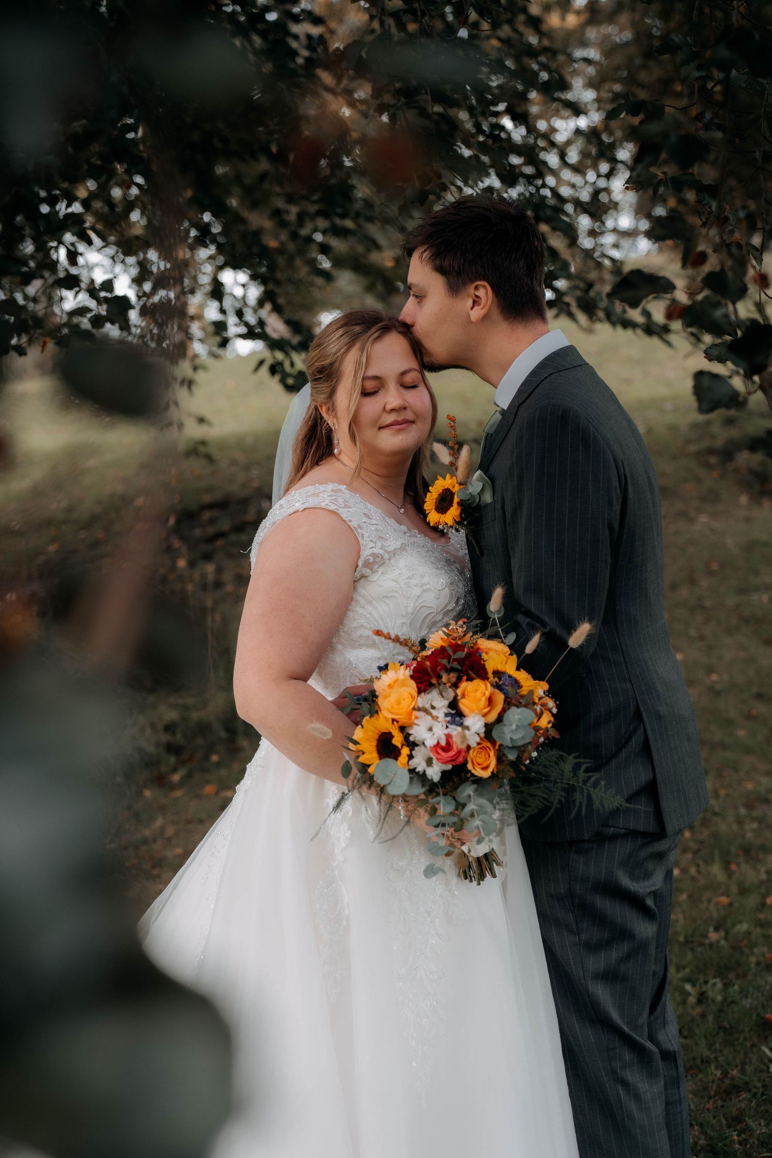 Bride holding autumn bouquet with sunflowers and roses, groom kissing her temple, outdoor fall wedding portrait.