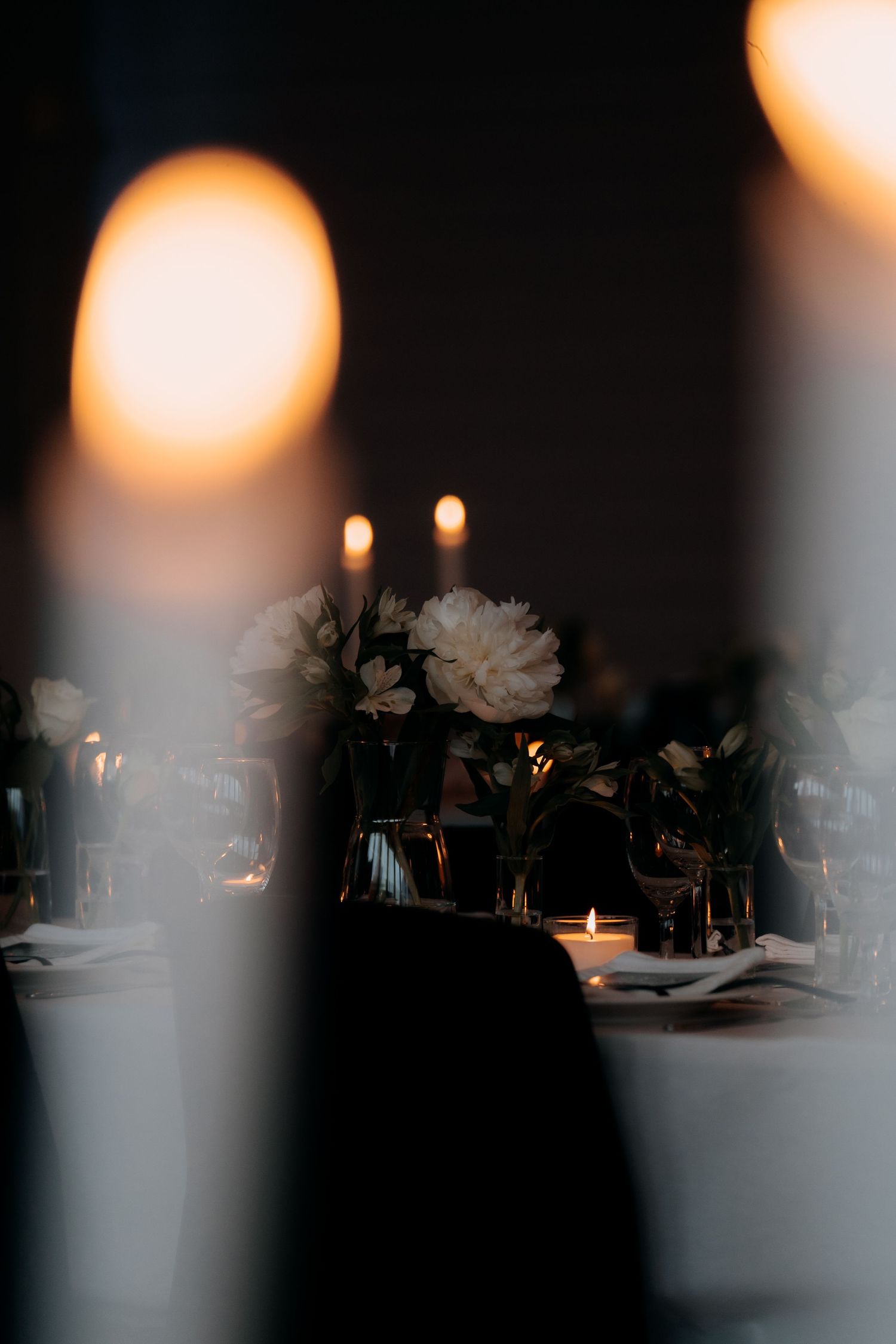 Elegant candlelit dinner table with white peony floral centerpieces and soft bokeh candlelight in a dark romantic setting.