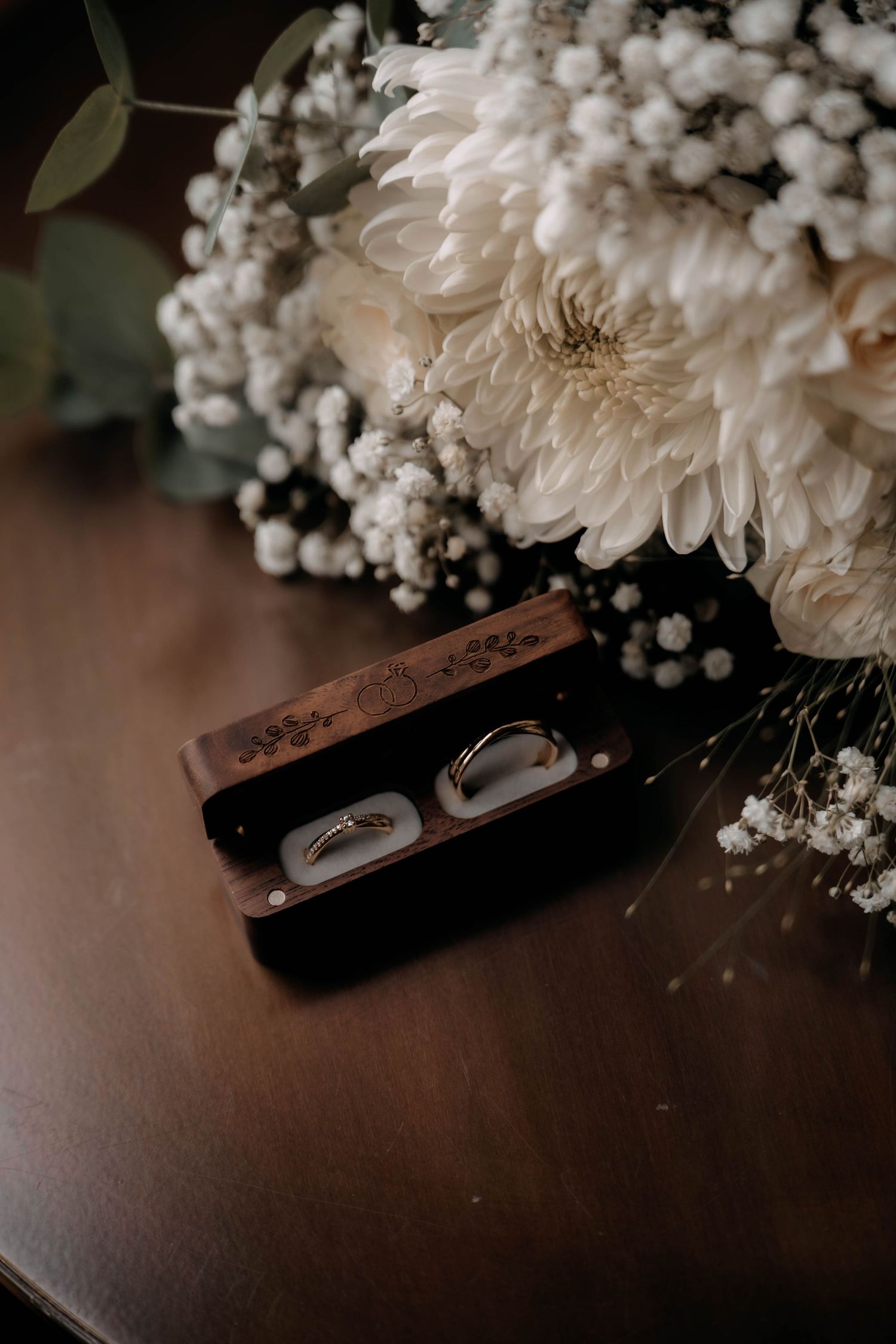 Two gold wedding rings in a wooden box beside a white dahlia and baby's breath bridal bouquet on a dark wood table.