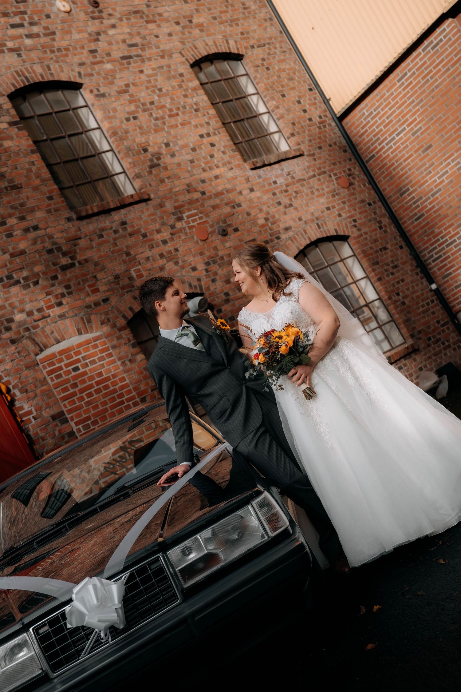 Bride and groom share a romantic moment leaning against a vintage car decorated with white ribbons outside a brick building.