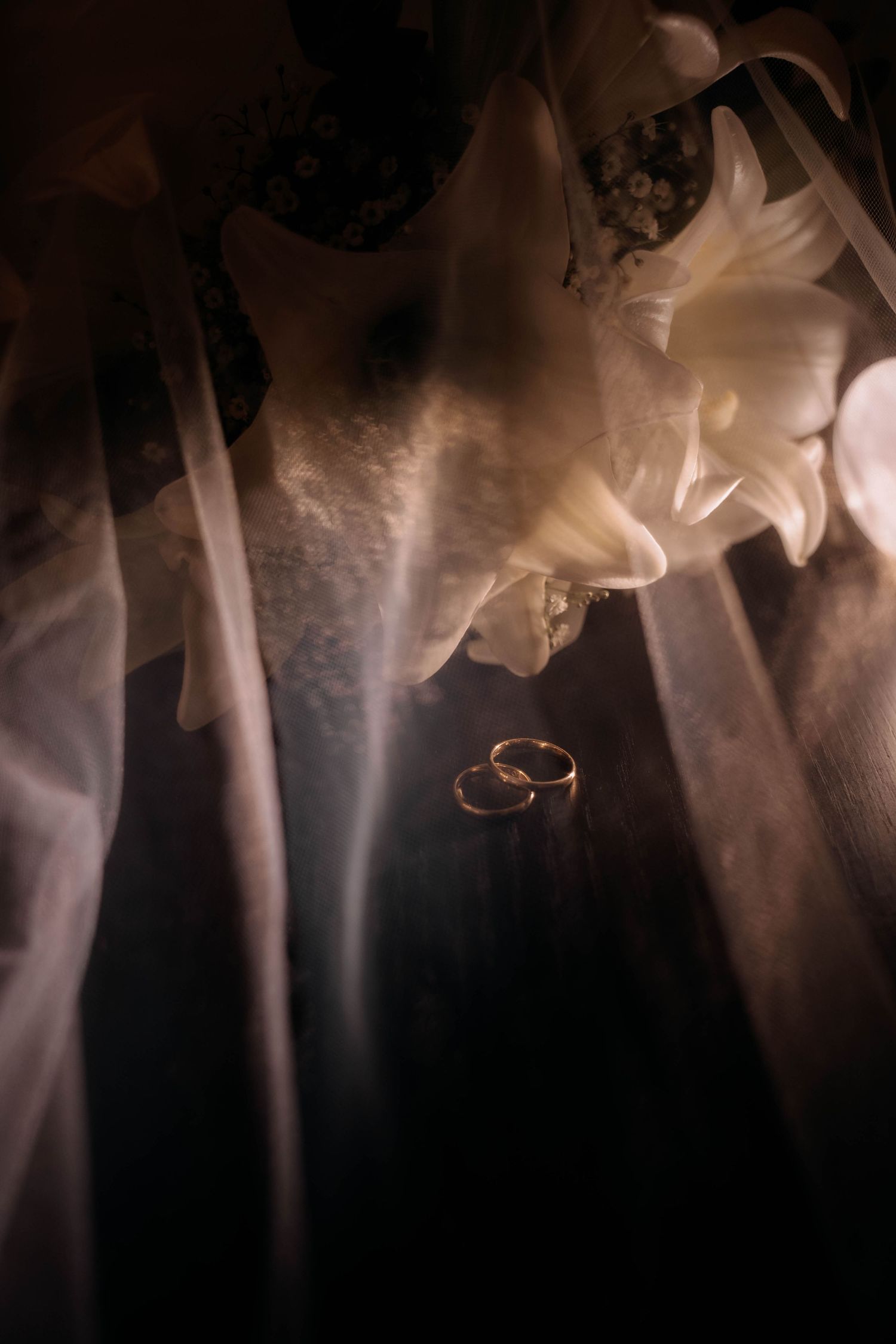 Two gold wedding rings rest beneath a bridal veil and white flowers in a dark, romantic setting.