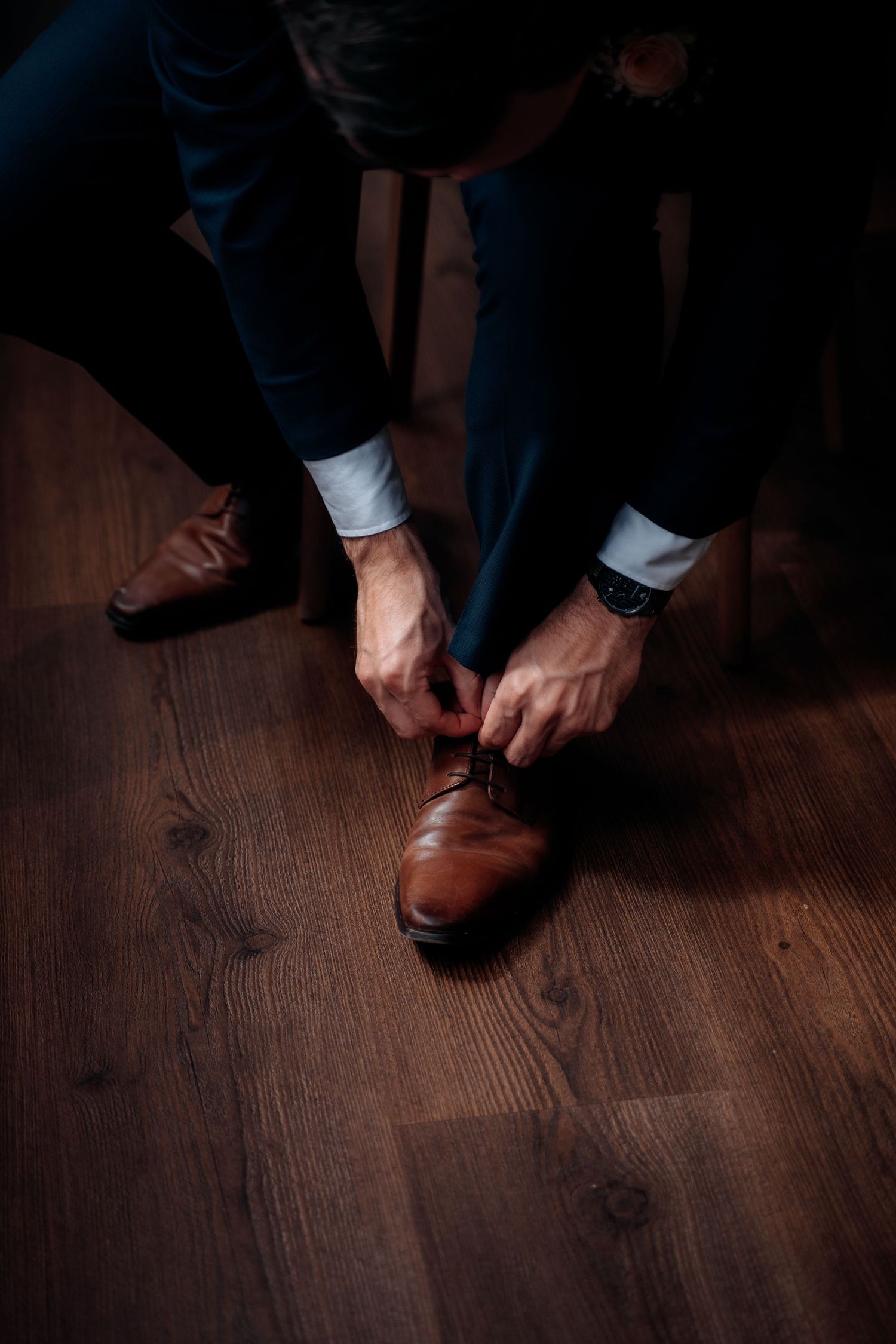 Man in navy suit tying brown leather Oxford dress shoes on wooden floor, wearing a watch.
